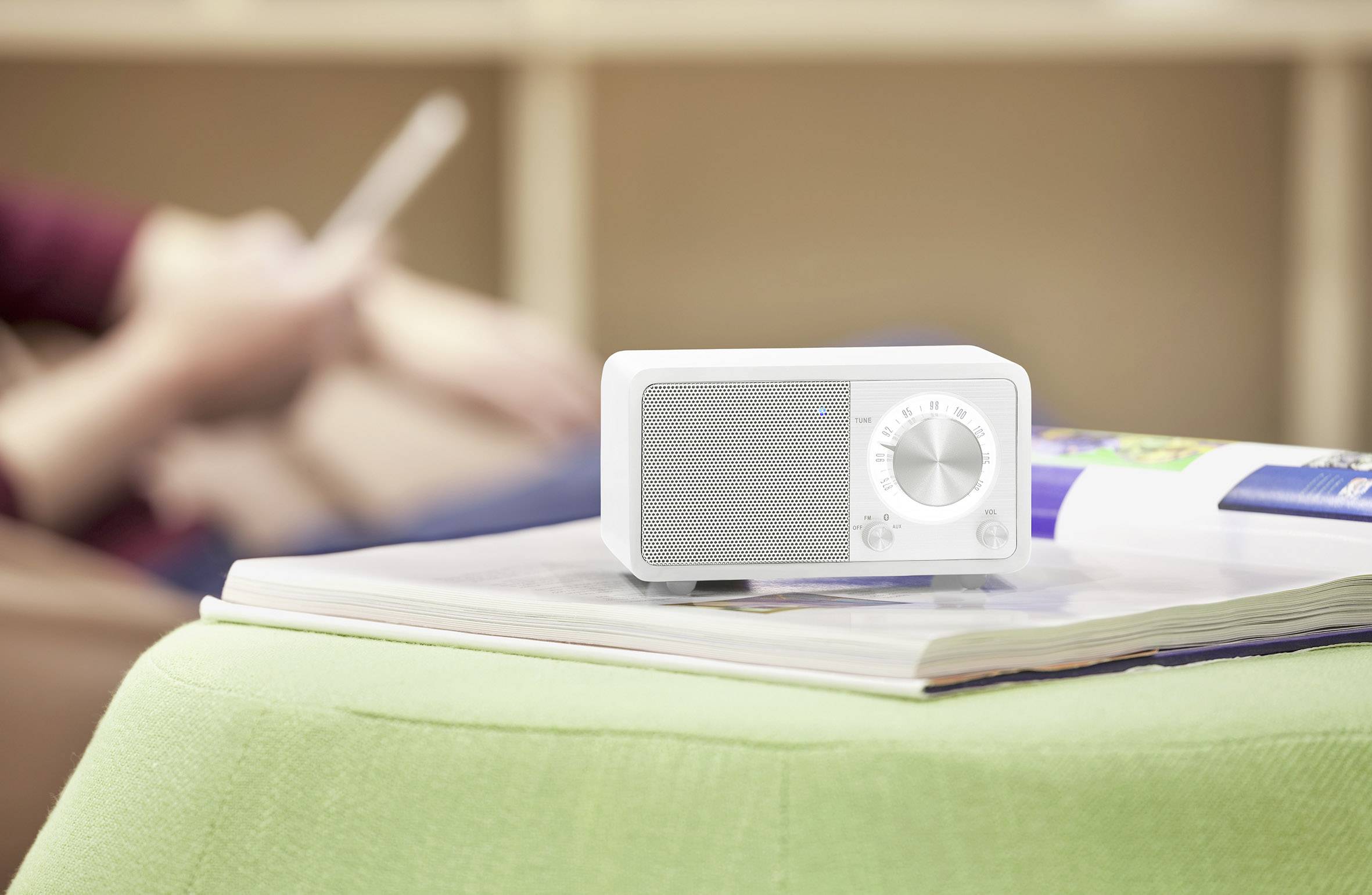 Een wit radio staat op een tafel met een groen kleed en opengeslagen boeken, op de achtergrond zit iemand met een mobiel apparaat.