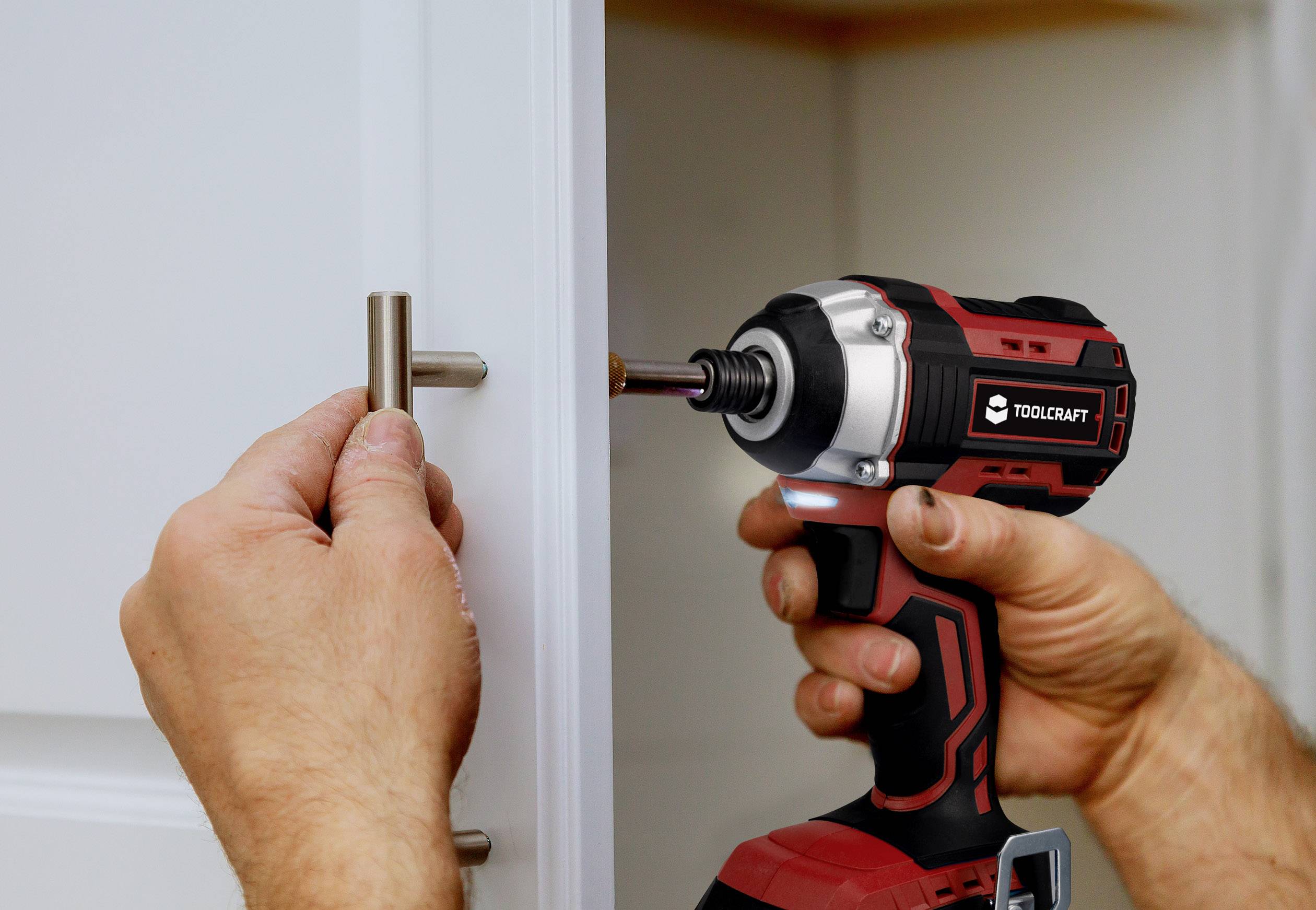 Een persoon monteert een deurgreep met een rode accuschroevendraaier. De greep wordt bevestigd aan een witte keukenkast.