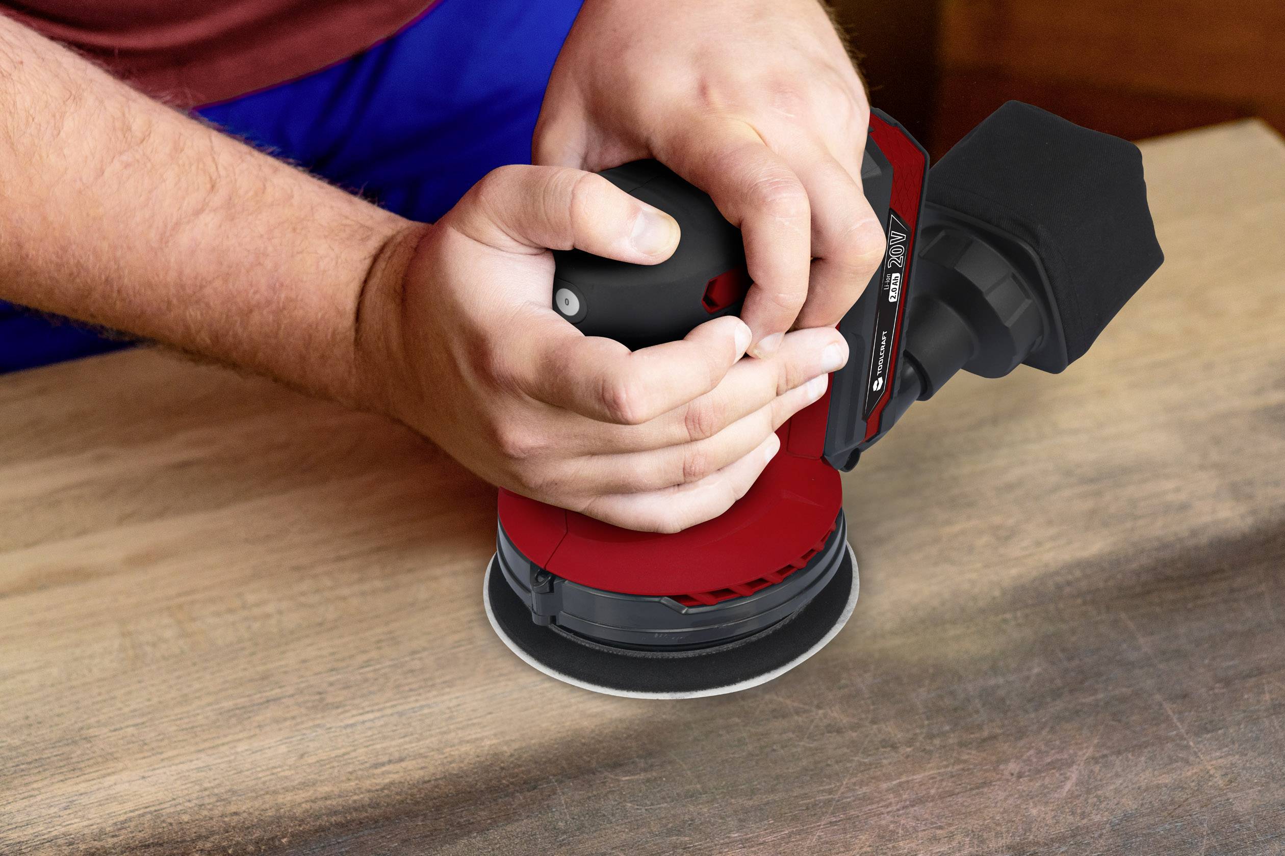 Een persoon slijpt een houten oppervlak met een elektrische slijpmachine. Het apparaat heeft een rode beschermkap.