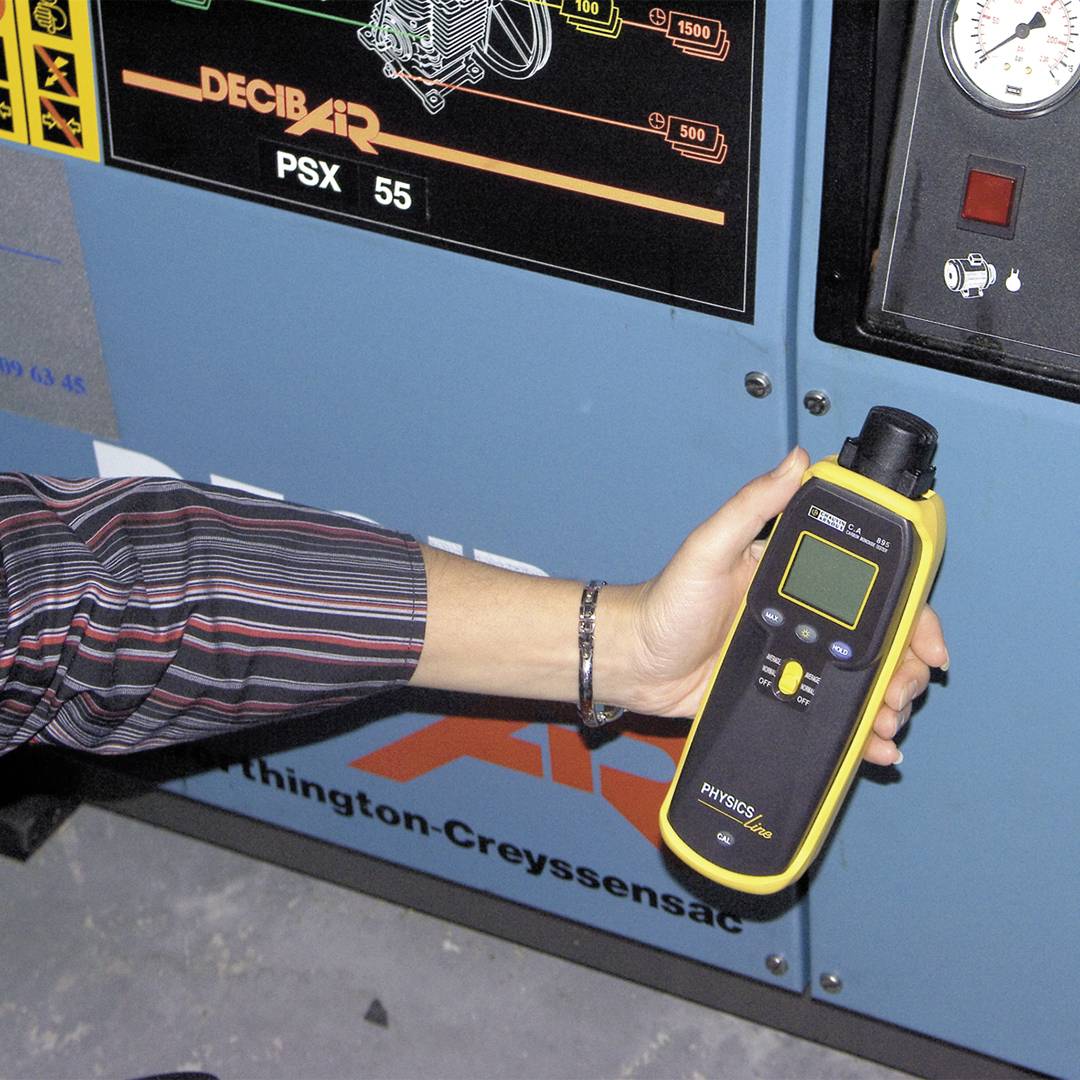 Een persoon meet met een geel handapparaat de druk aan een compressor, naast een manometer en het opschrift 'DECIBAIR PSX 55'.