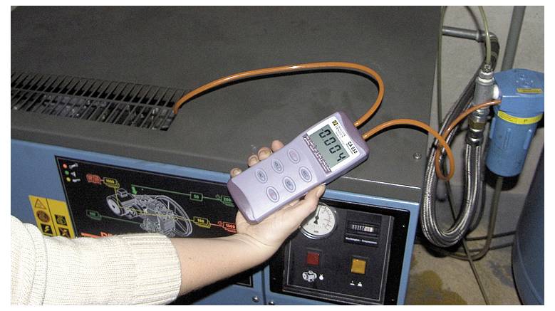 Een persoon houdt een meetapparaat vast dat de waarde '0.004' weergeeft, voor een elektrisch apparaat met een oranje kabel in een industriële omgeving.