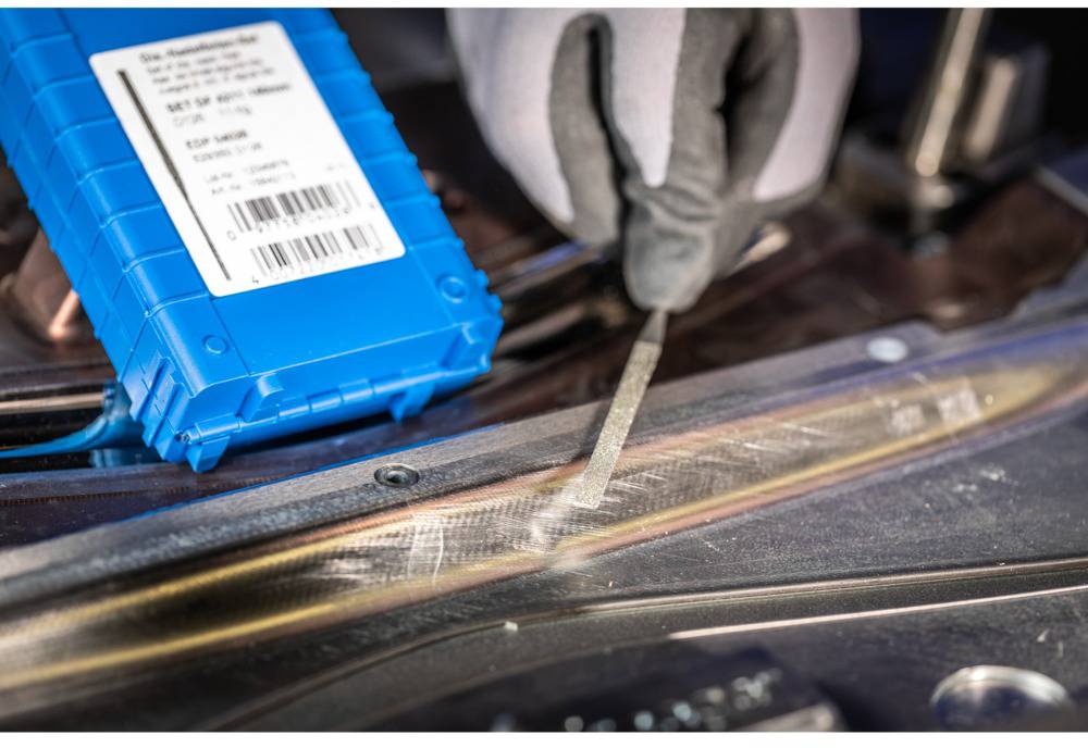 Hand die een vijl gebruikt op een metalen sjabloon, naast een blauw object met een etiket. Scène toont nauwkeurig ambachtelijk werk.