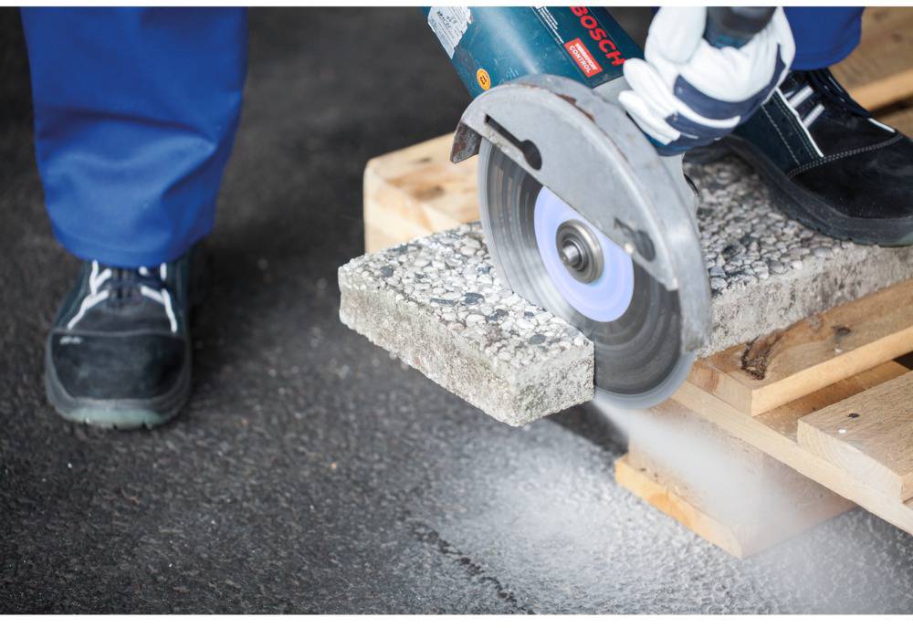Persoon snijdt betonplaat met haakse slijper, stofwolk zichtbaar. Betonplaat ligt op houten pallet. Veiligheidsschoenen gedragen.