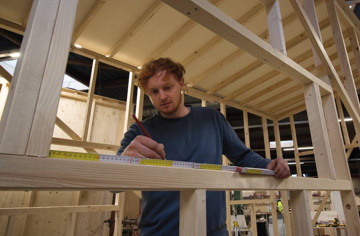 Een man meet met een meetlint en noteert op een houten frame in een werkplaats, geconcentreerd aan het werk aan een houtbouwproject.