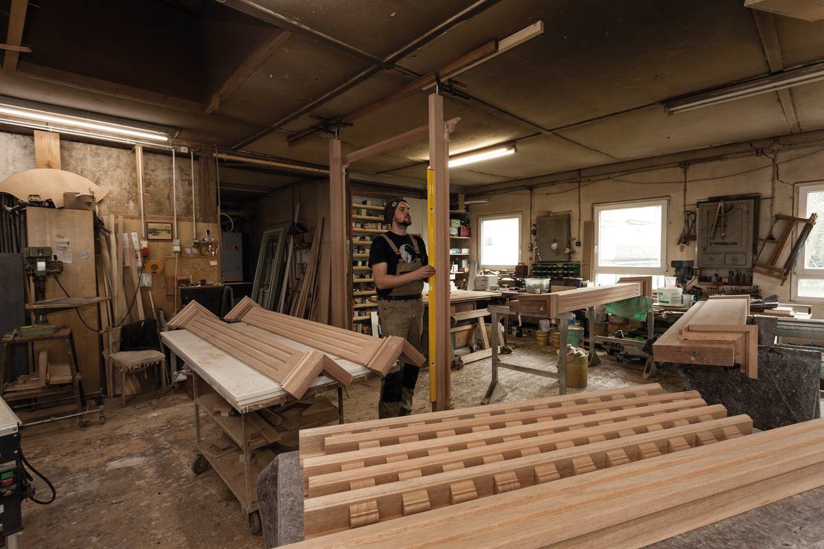 Een werkplaats met een ambachtsman die houtbewerkingen uitvoert. Er zijn verschillende houten stukken, gereedschappen en machines te zien.