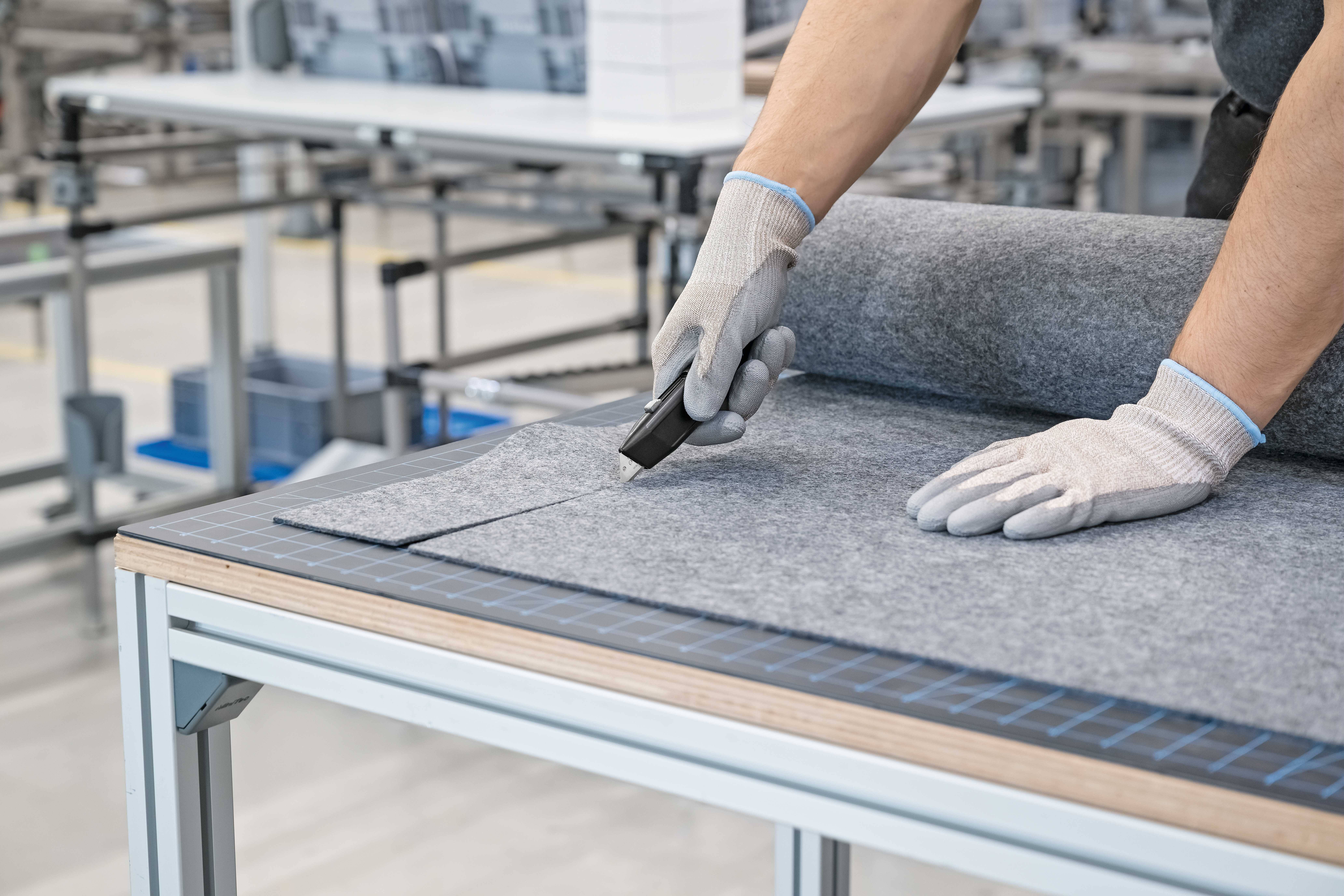 Een persoon snijdt een grijs stoffen materiaal met een cuttermes op een tafel. De achtergrond toont een fabriekromgeving.