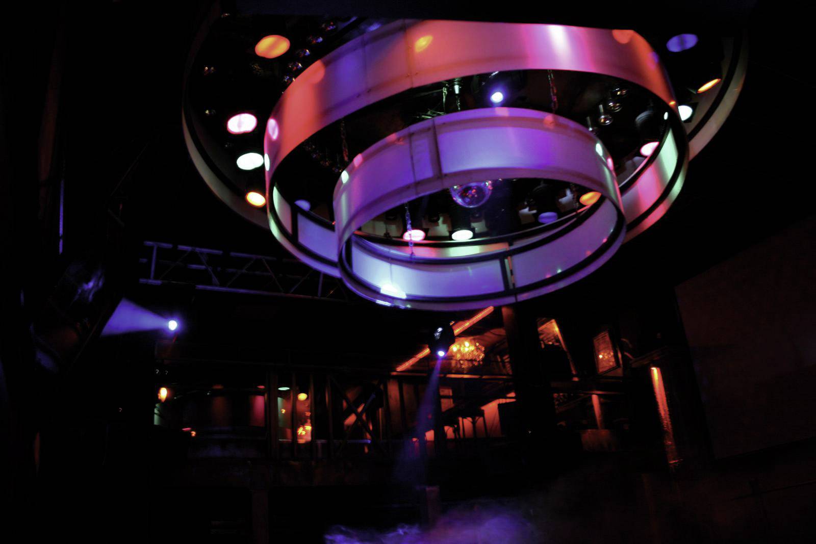 Een club met kleurrijke verlichting. Op de voorgrond hangen felgekleurde lichtcirkels van het plafond. De sfeer is donker en sfeervol.