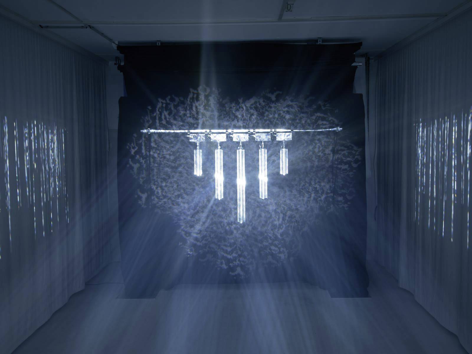 Een kunstwerk met verticaal hangende, verlichte buizen aan het plafond. Blauwe lichtstralen en schaduwen projecteren patronen.