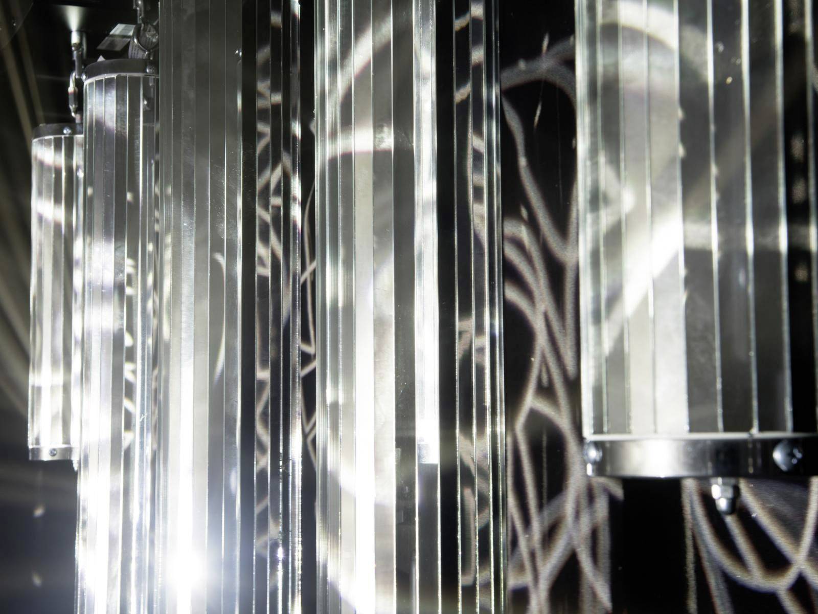 Metalen, verticale cilinders werpen reflecterende lichtpatronen op een donker oppervlak. De scène oogt abstract en futuristisch.