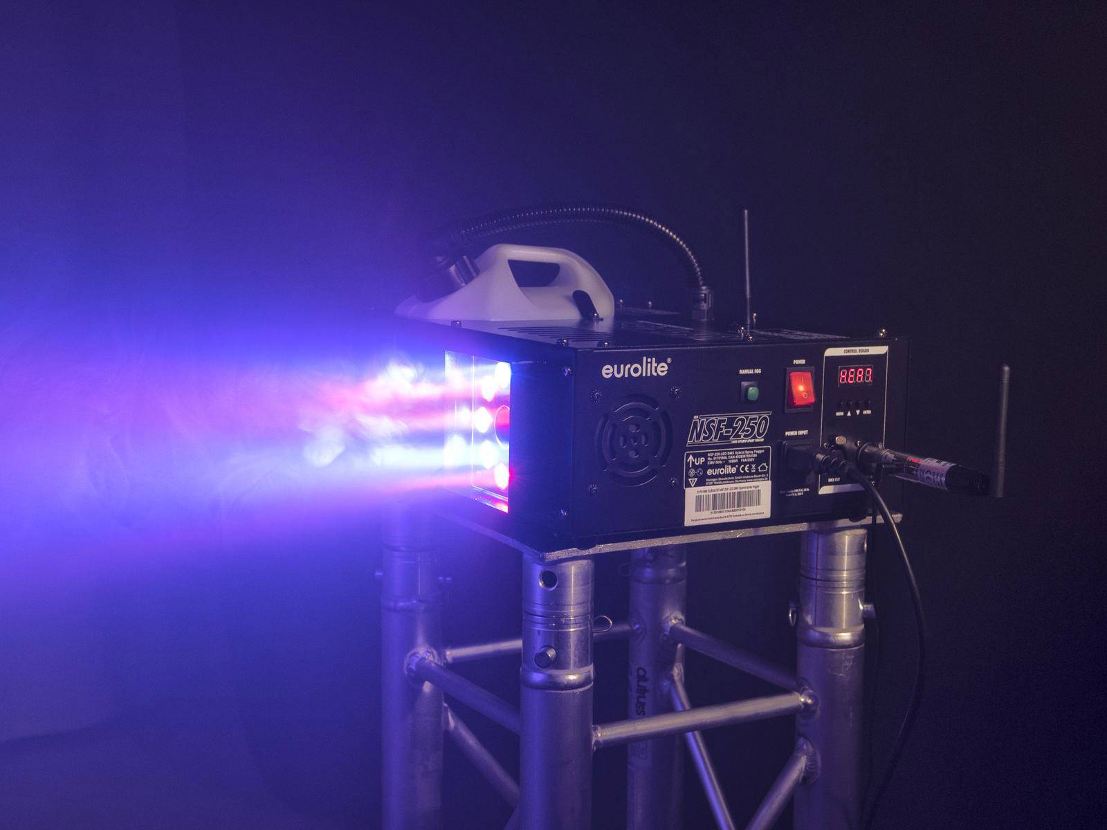 Rookmachine op een stalen frame geeft gekleurde lichten en rookstraal af, omgeven door een donkere achtergrond.