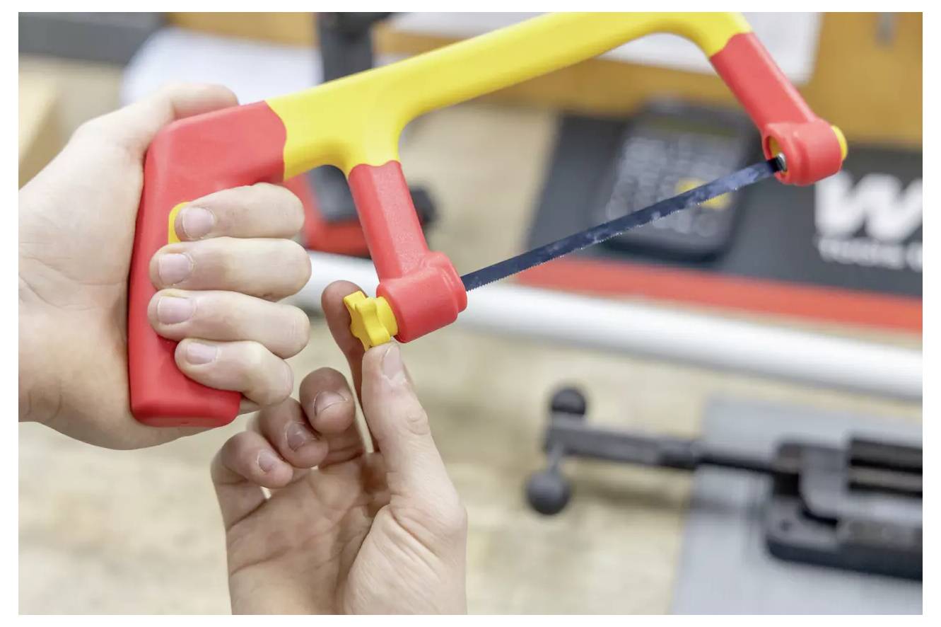 Een persoon stelt de spanning van een kleine handhaakzaag met een rood en geel handvat in, geplaatst op een werkbank met onscherpe gereedschappen op de achtergrond.