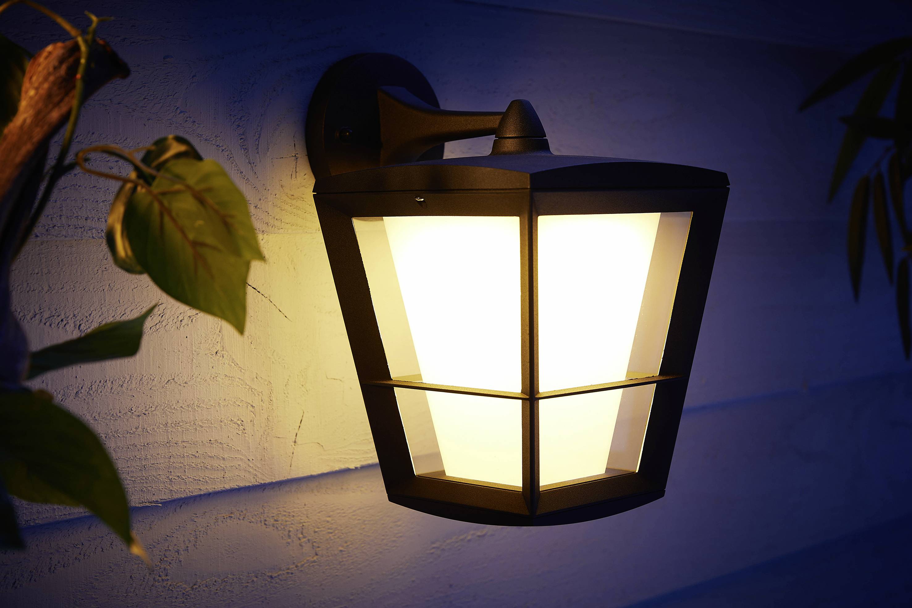 Buitenmuurlamp die in het donker schijnt, gemonteerd aan een houten gevel met enkele bladeren op de voorgrond.