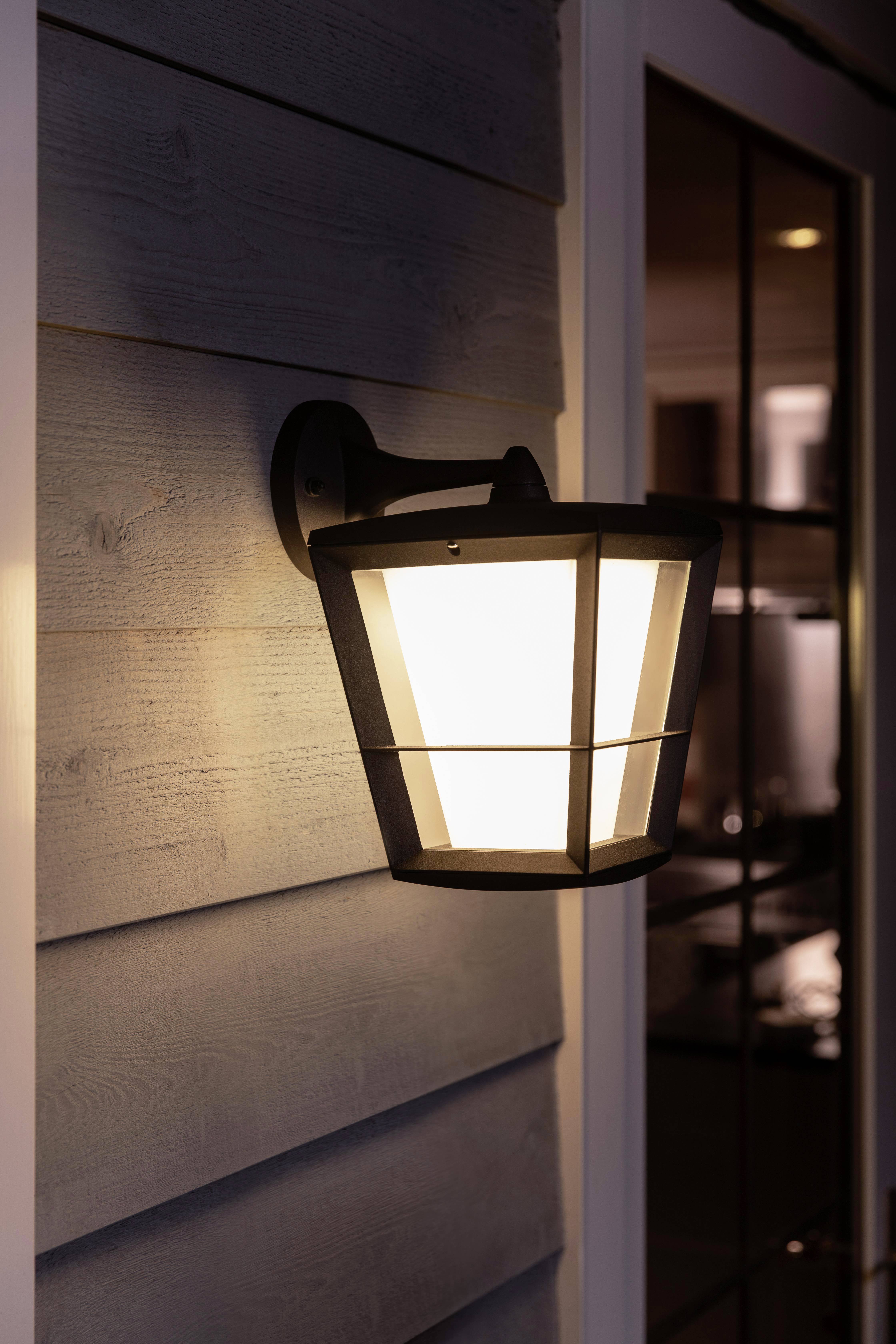 Buitenlamp verlicht een veranda bij het invallen van de avond, gemonteerd aan een muur met houten bekleding, naast een glazen deur.