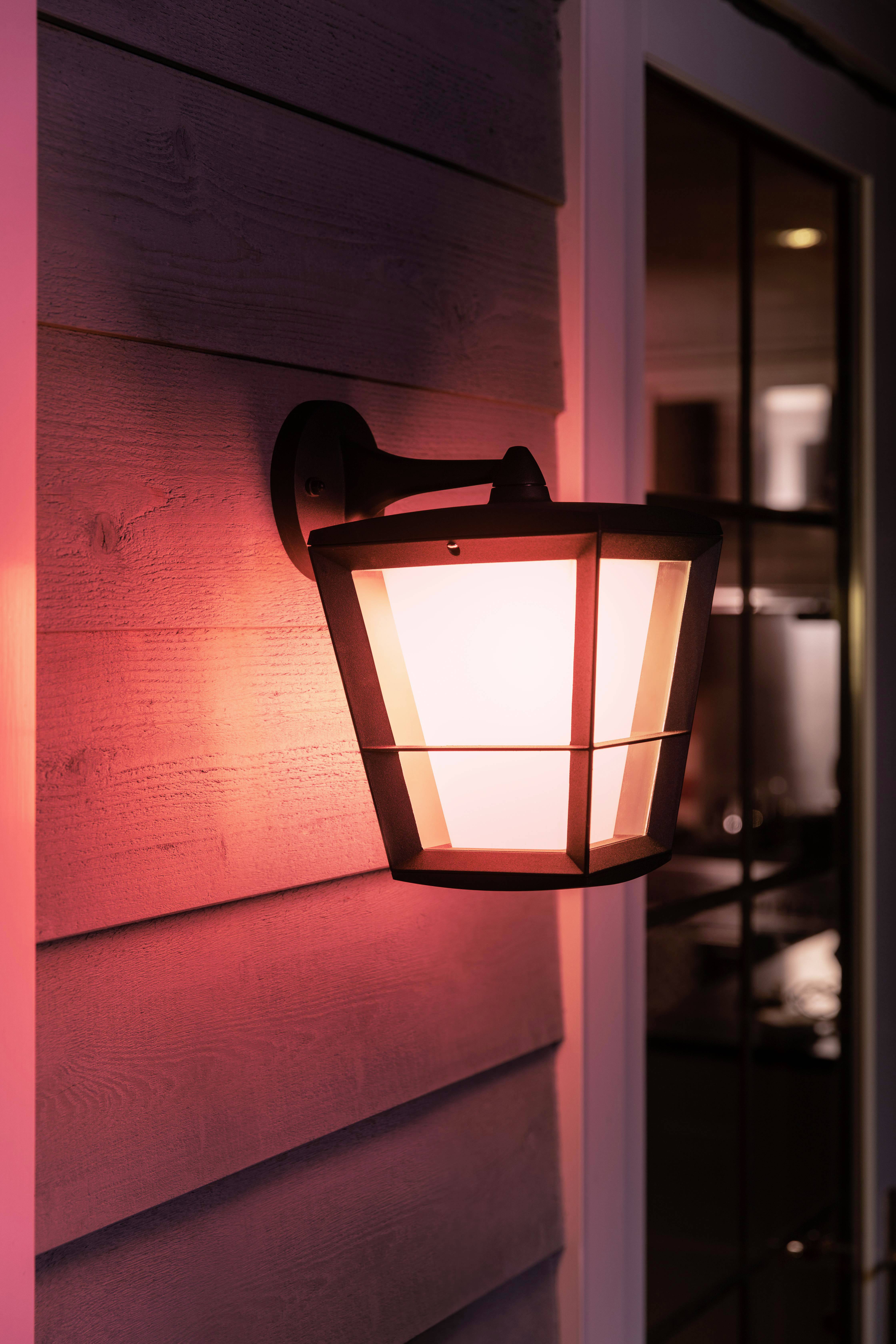 Buitenmuurlamp verlicht muur in zacht rood. Lampvorm modern, rechthoekige ramen weerkaatsen licht, creëren een gezellige avondsfeer.