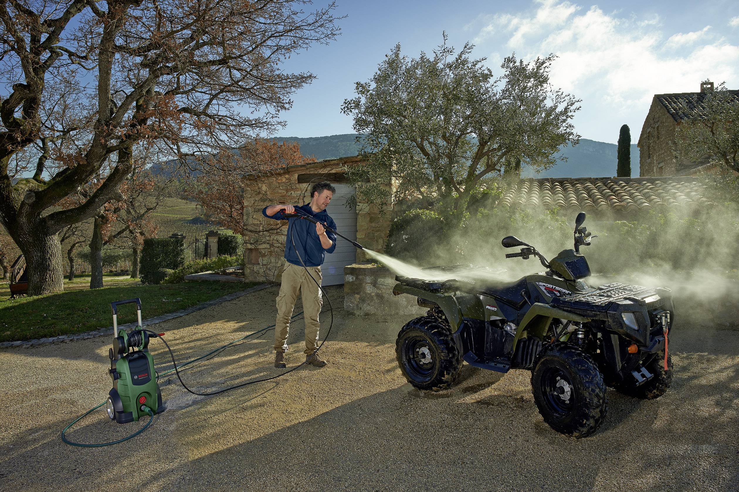 'Een man reinigt een ATV met een hogedrukreiniger op een erf. Bomen en een gebouw zijn op de achtergrond zichtbaar.'