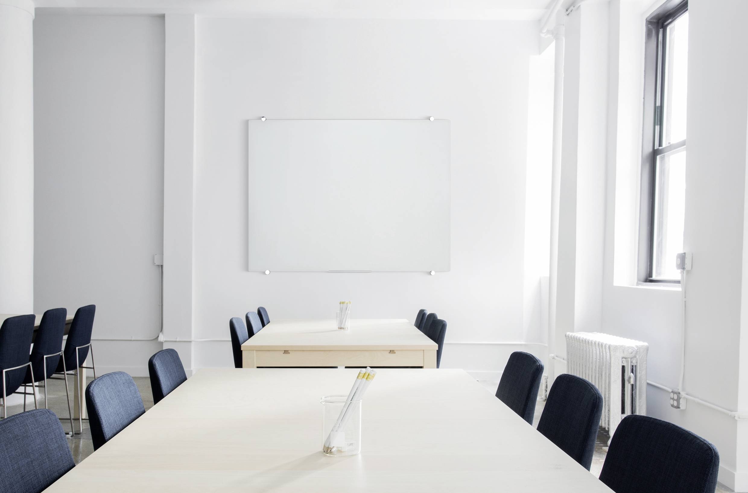 Een moderne, lichte vergaderzaal met twee tafels, zwarte stoelen en een leeg whiteboard aan de witte muur.