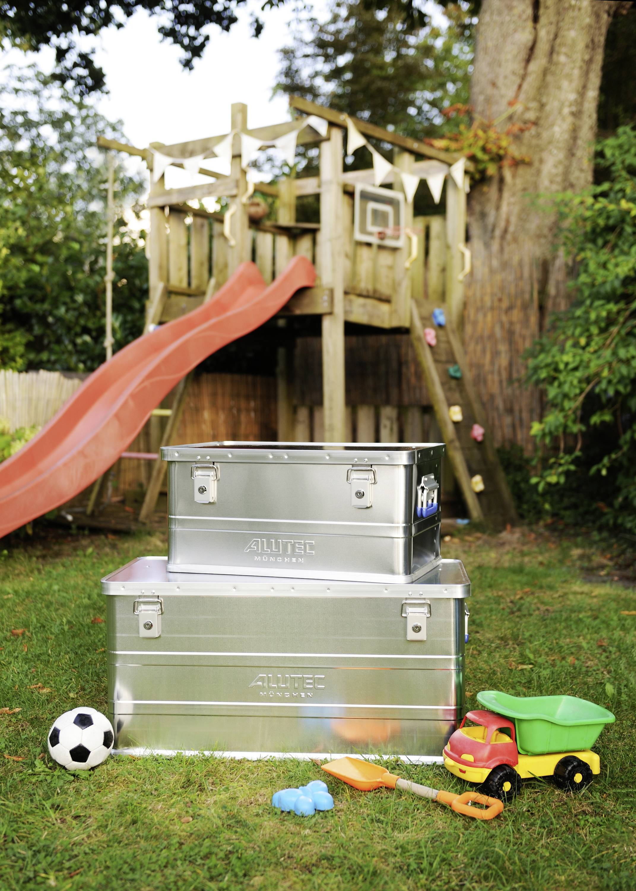 Twee zilveren dozen staan in de tuin voor een klimrek met glijbaan; ernaast een voetbal, speelgoedauto en schepjes.