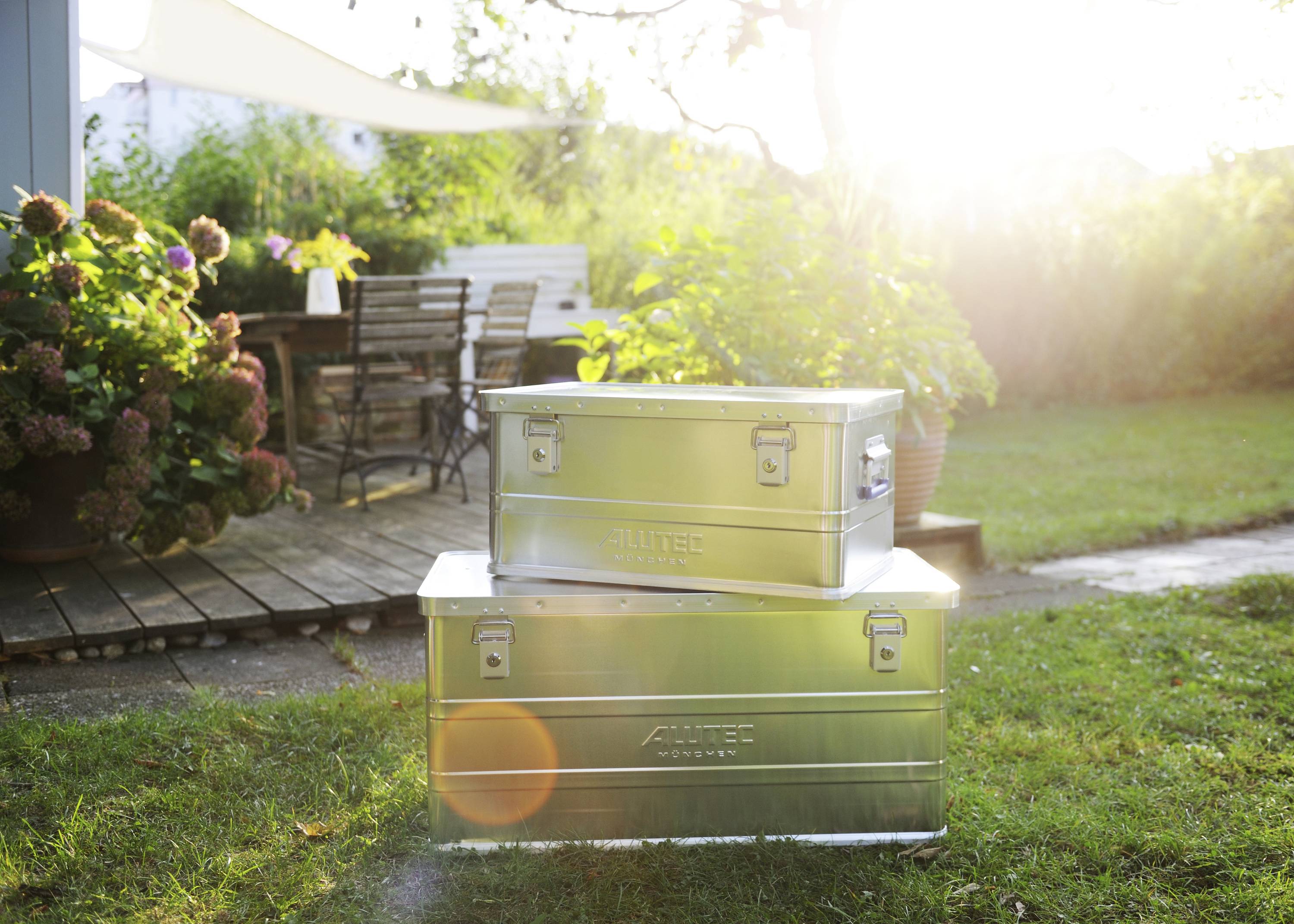 Twee metalen dozen staan op het gazon in de tuin, de zon schijnt helder op de achtergrond. Tuinmeubilair is zichtbaar op de achtergrond.