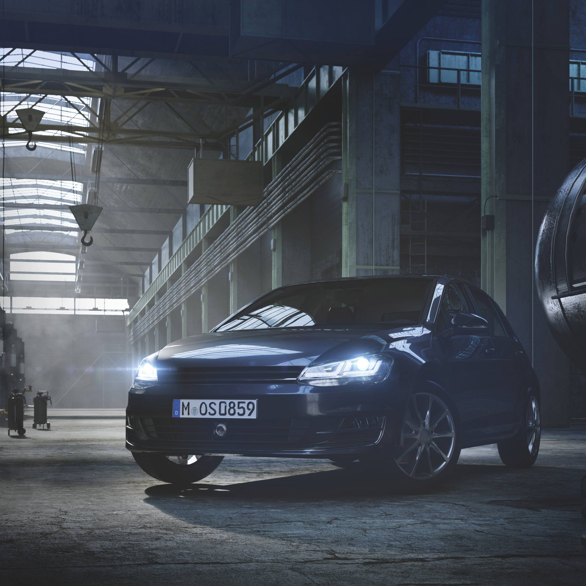 Een blauwe auto staat in een verlichte, industriële hal met hoge plafonds en grote ramen. Links zijn gereedschappen te zien.