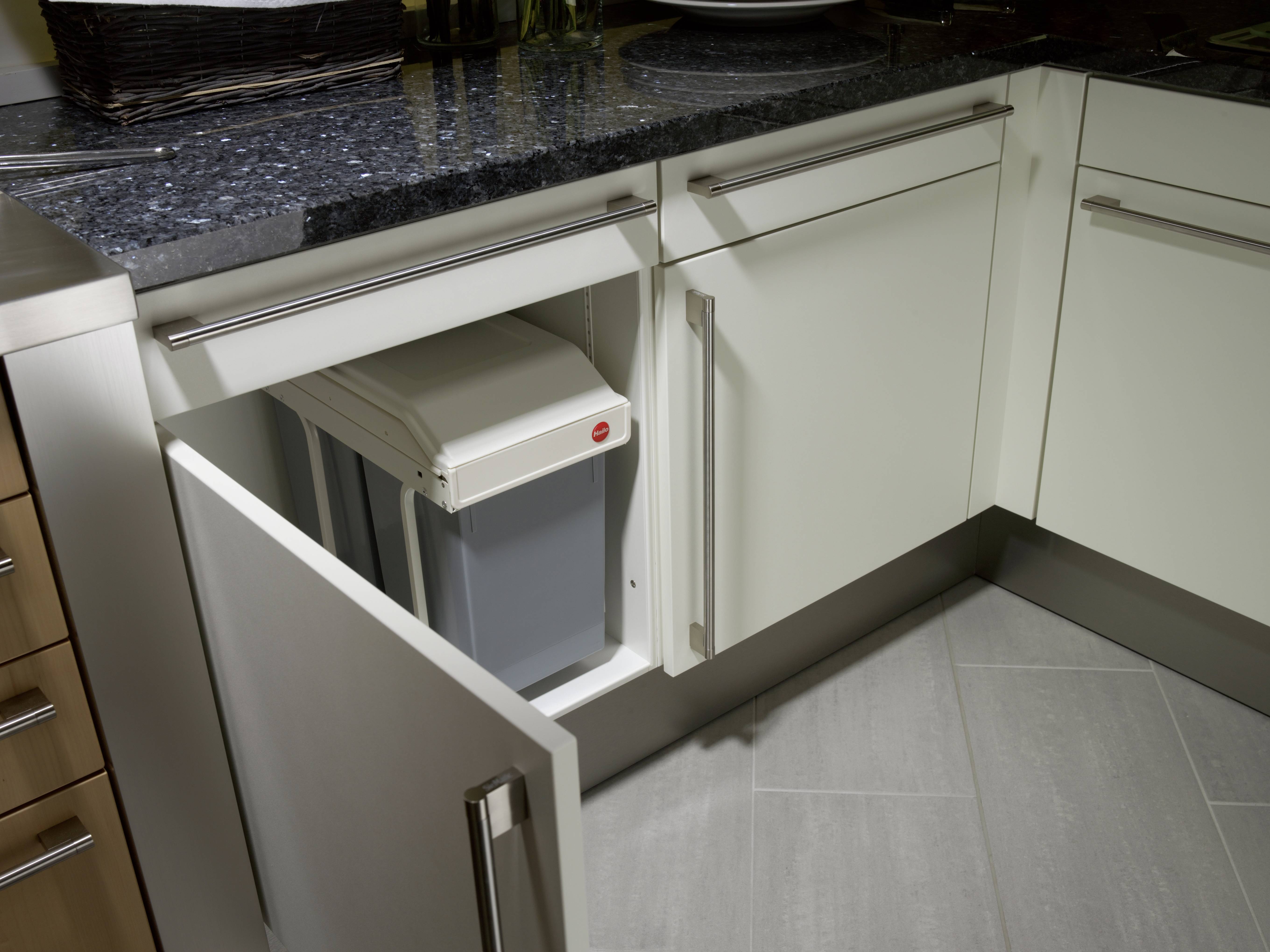 Onderkast in een moderne keuken met een open kastdeur waarachter een grijze vuilnisemmer zichtbaar is. Lichte kasten en een donker granieten werkblad.