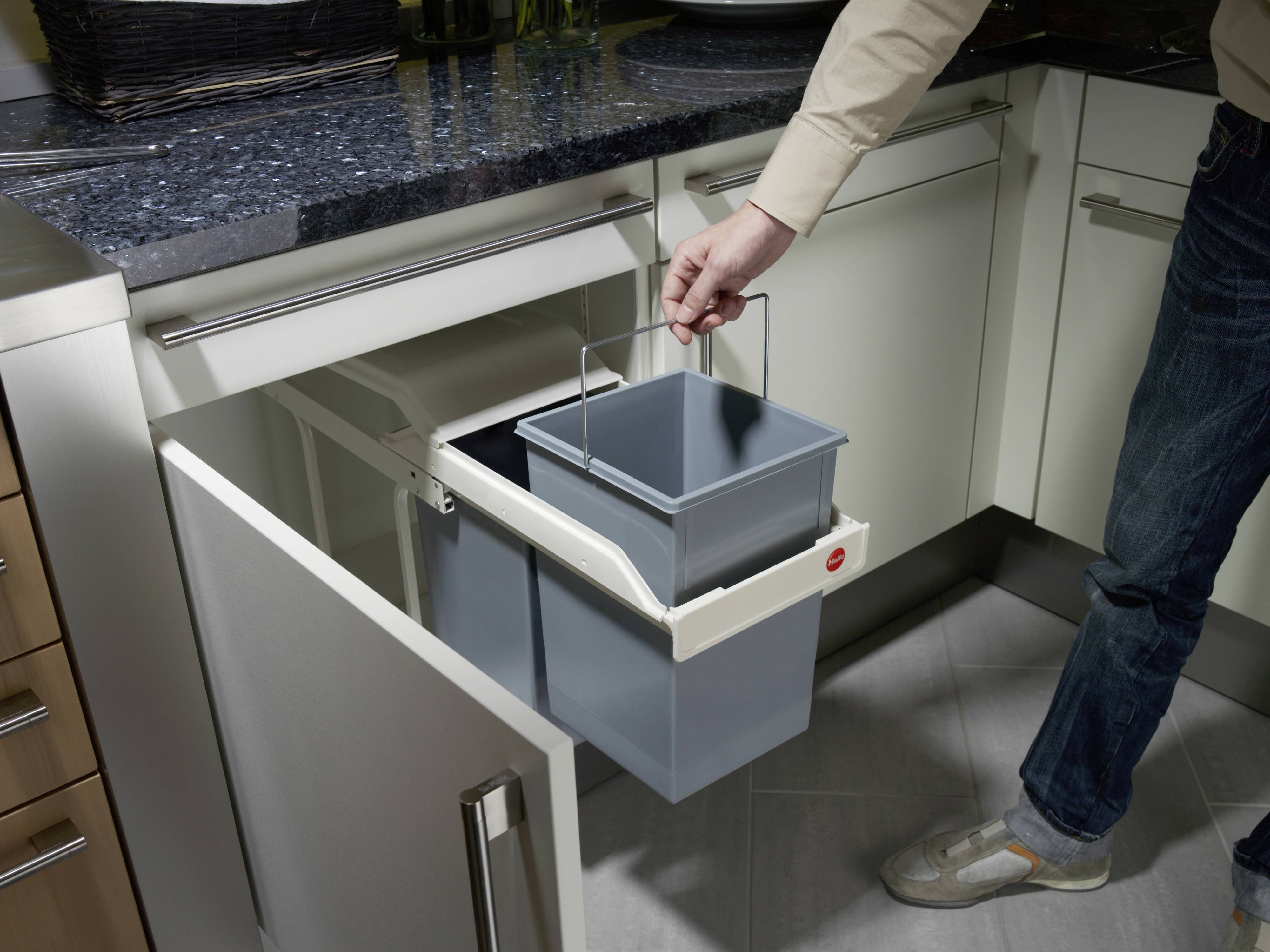 Een persoon opent een kast onder de gootsteen en tilt een uitneembare prullenbak uit een afvalscheidingssysteem.