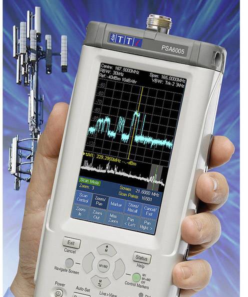 Een handapparaat voor frequentieanalyse wordt getoond. Het scherm toont een spectrum. Op de achtergrond is een zendmast zichtbaar.
