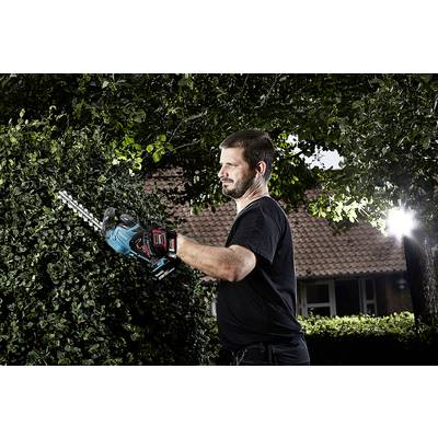 Een man snoeit een groene haag met een elektrische heggenschaar, omringd door bomen. Op de achtergrond een bakstenen gebouw.