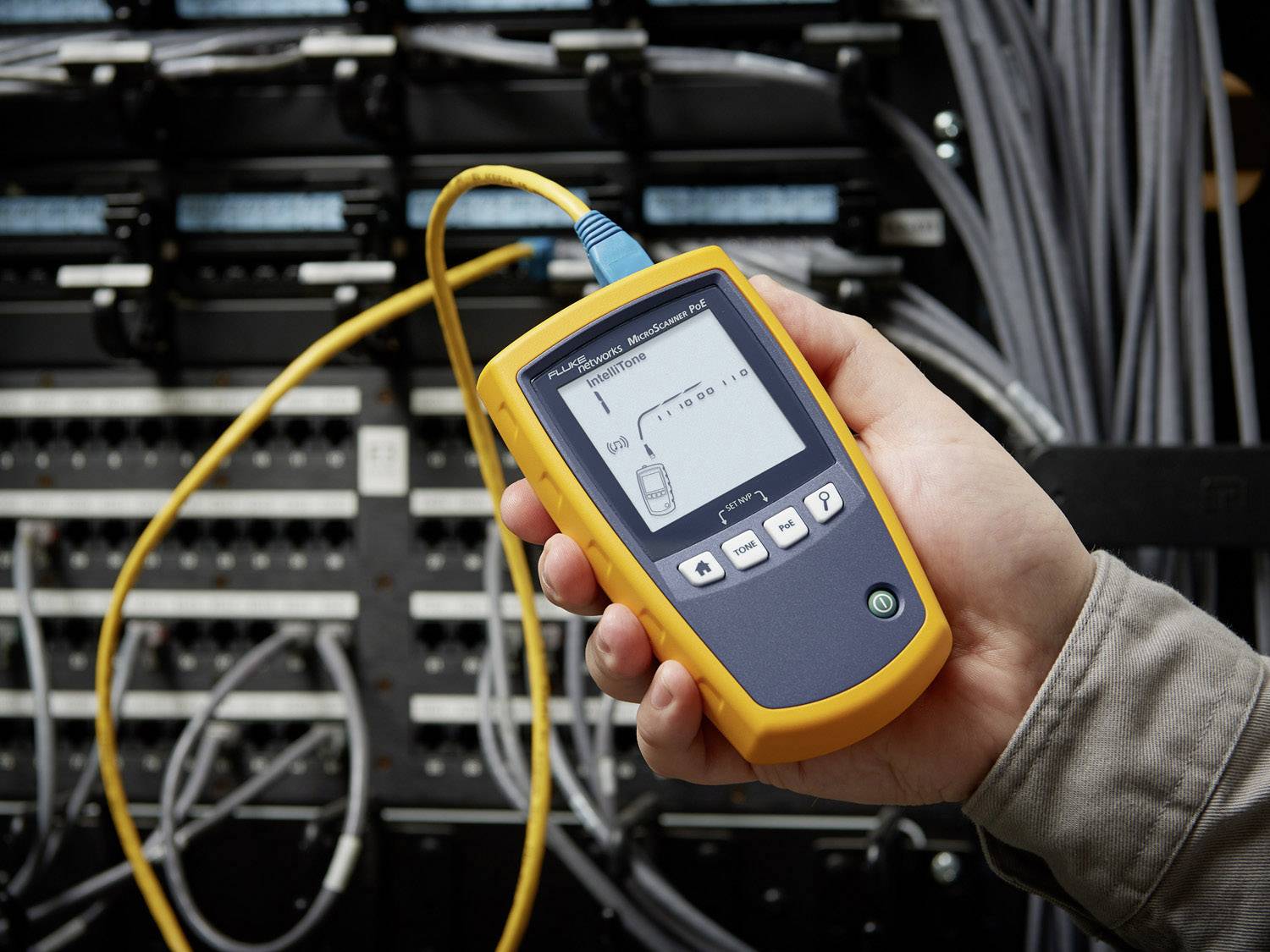 Een hand houdt een netwerkkabeltester voor een serverrek. De tester toont meetwaarden op het display. Kabels zijn aangesloten.