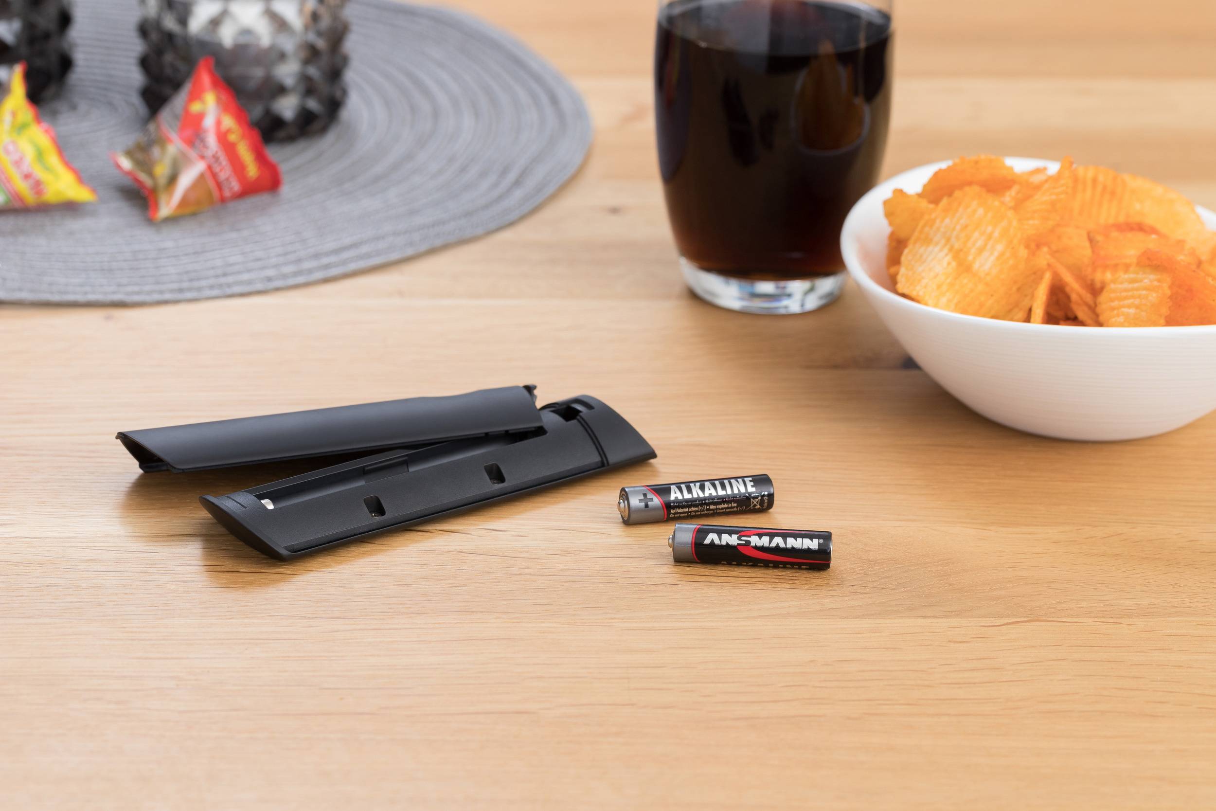 Een afstandsbediening zonder deksel en batterijen ligt naast een schaal met chips en een glas donkerrode vloeistof op tafel.