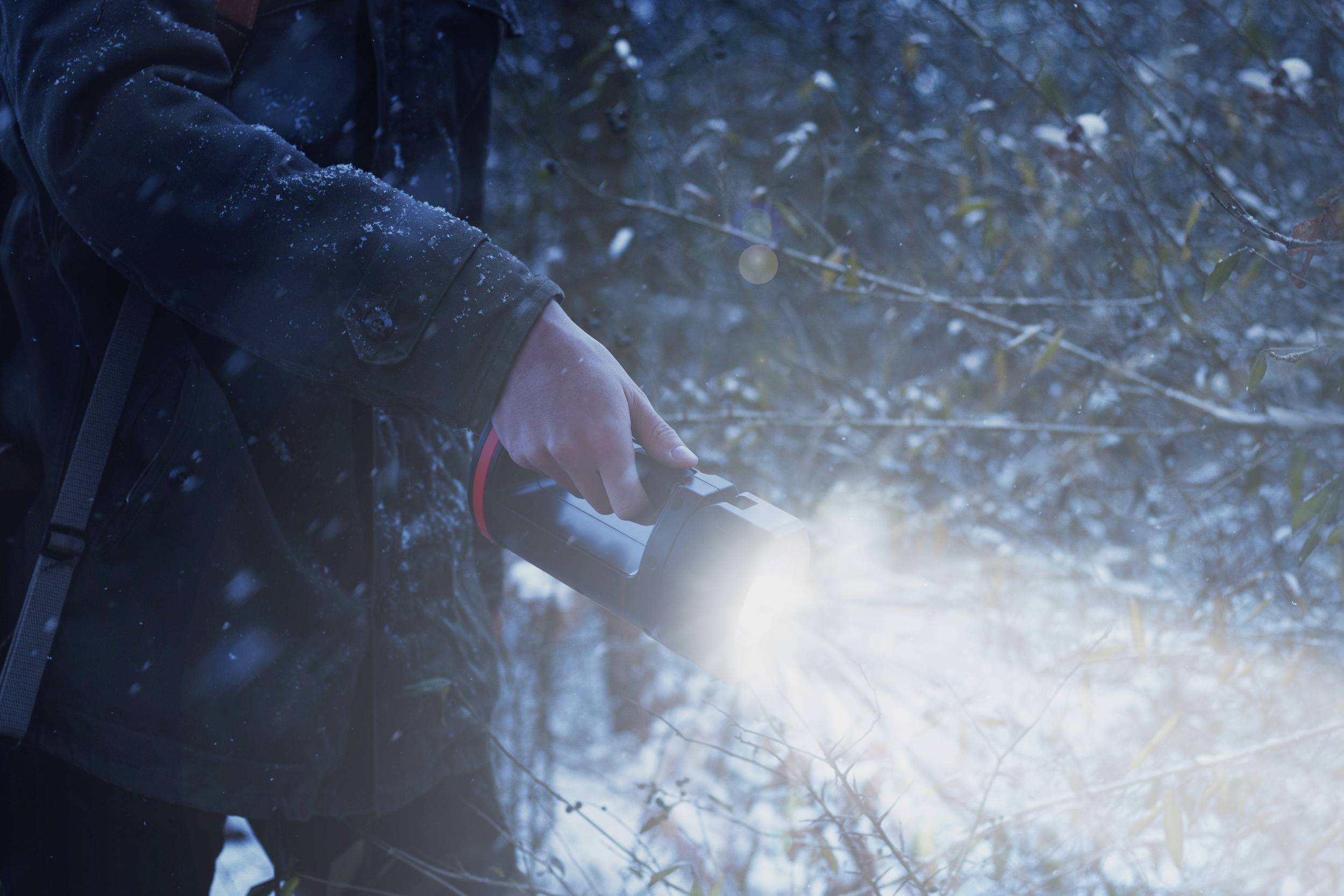 Een persoon houdt een felle zaklamp in een besneeuwde bos, terwijl sneeuwvlokken neerdalen.