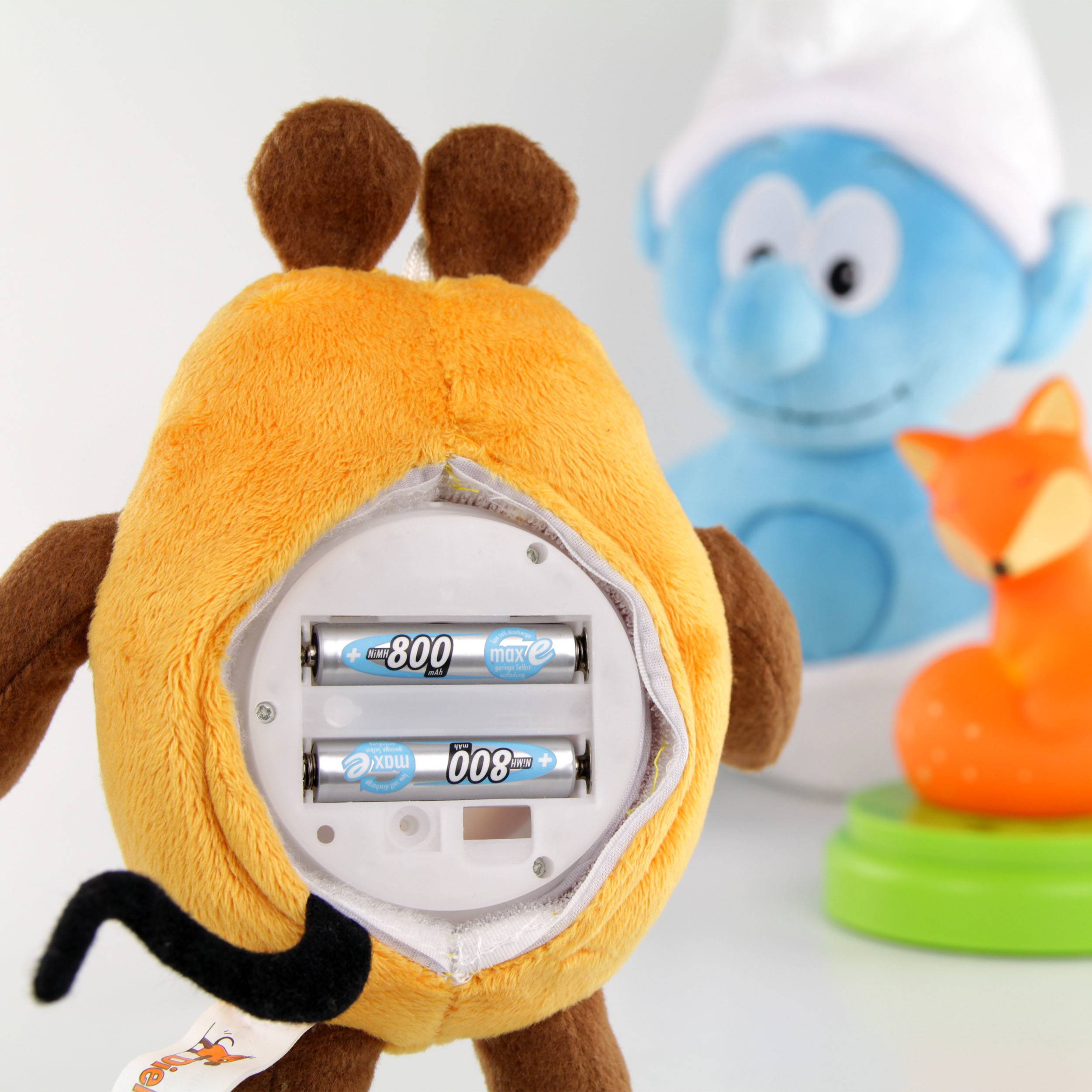 Een knuffeldier met batterijvak is geopend. Op de achtergrond zijn een blauw speelgoed en een oranje vos wazig te zien.