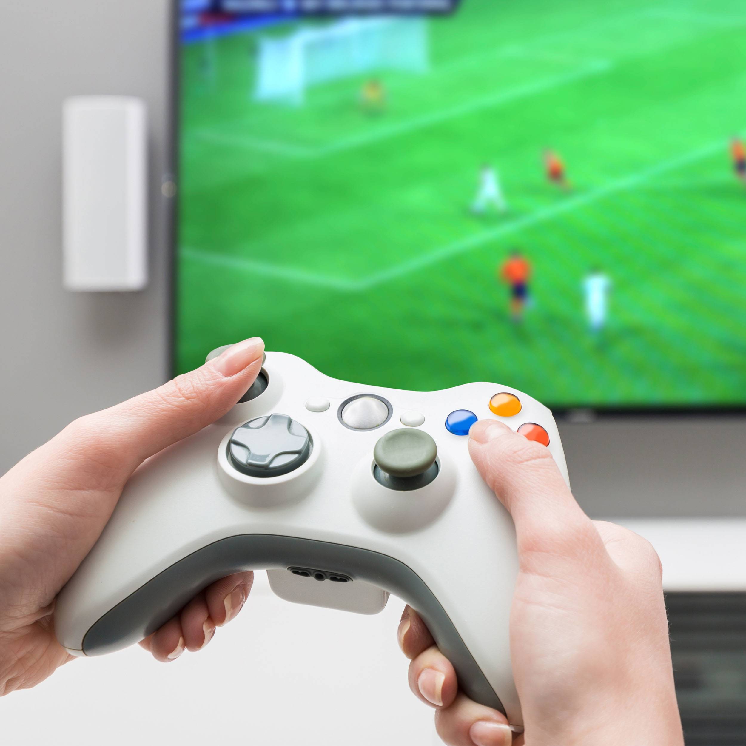 Een persoon houdt een videogame-controller vast voor een televisie waarop een voetbalspel wordt gespeeld. Handen op de voorgrond, beeldscherm op de achtergrond.