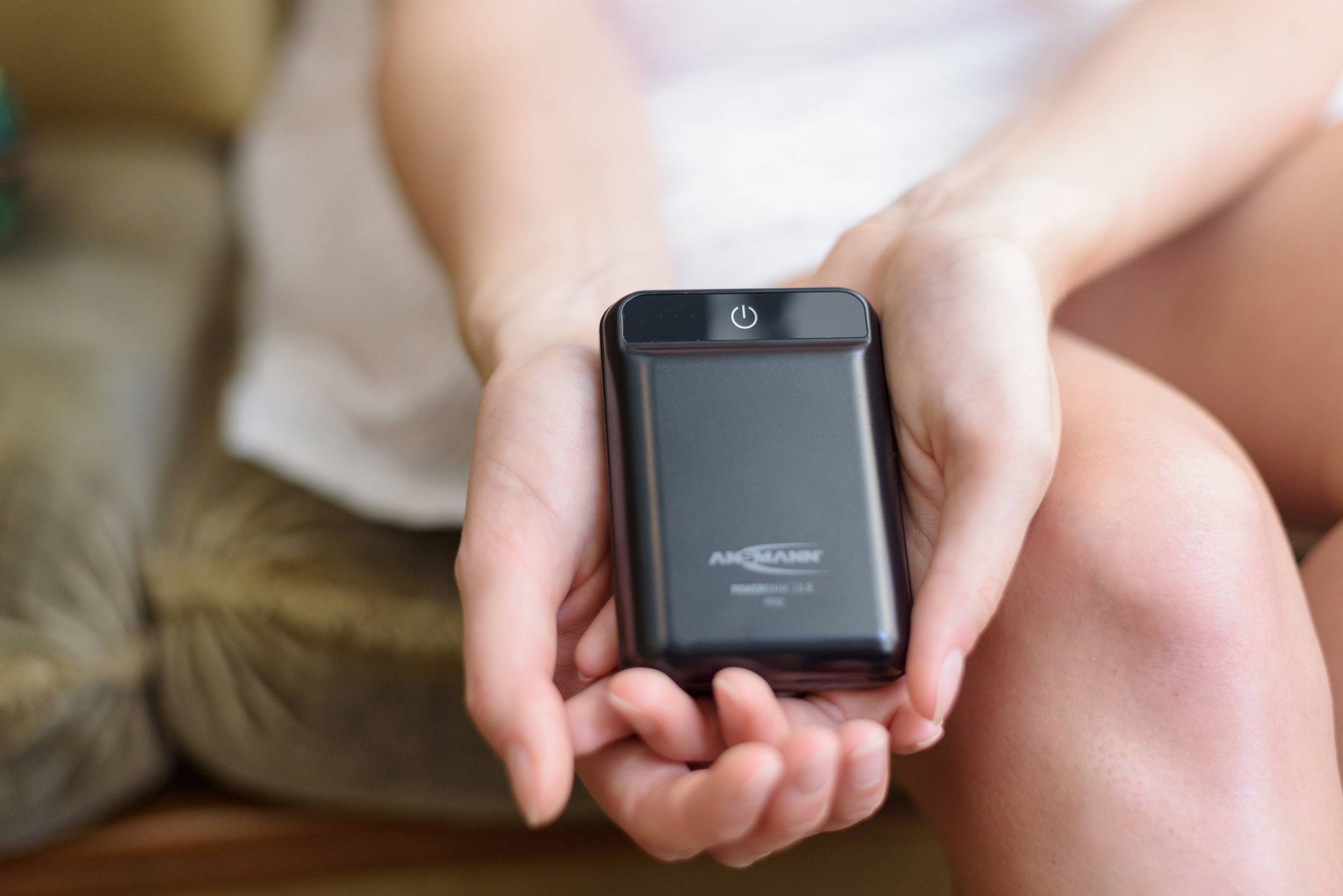 Een persoon houdt een kleine, zwarte powerbank in beide handen. Aan de bovenkant is de aan/uit-knop zichtbaar, terwijl hij zit op een bank of stoel.