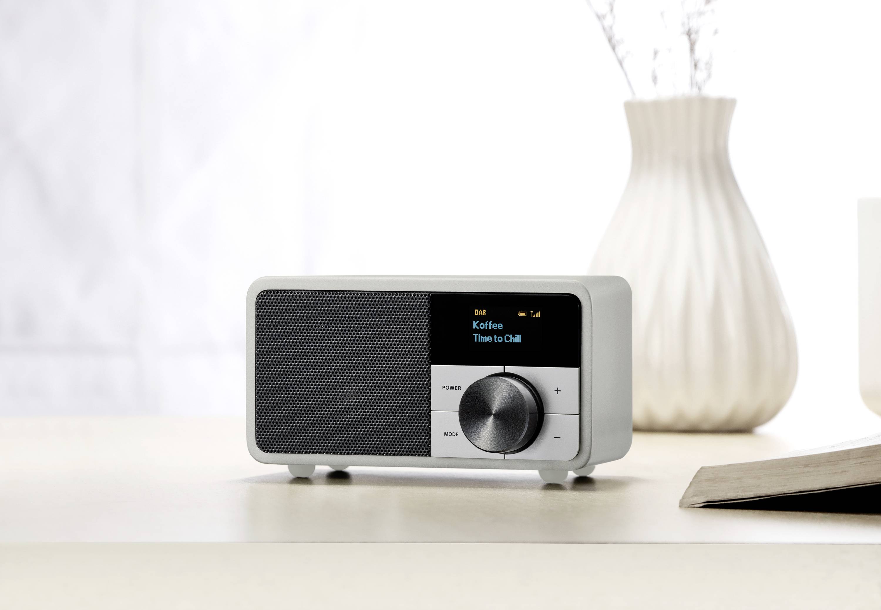 Een wit digitaal radio met een zwart scherm toont 'Kaffen' en 'Time to Chill'. Het staat op een tafel naast een boek en een vaas.