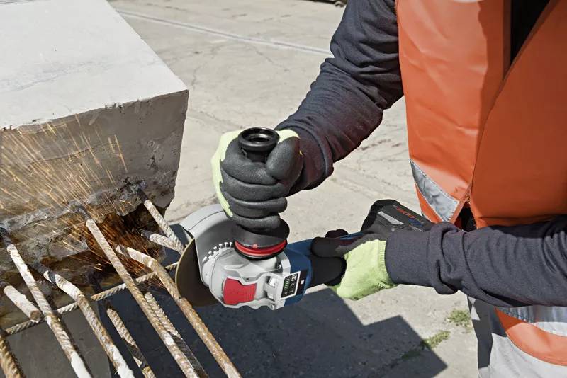Persoon in veiligheidshesje gebruikt een haakse slijper om metalen staven uit beton te snijden. Vonken spatten rond tijdens het werk.