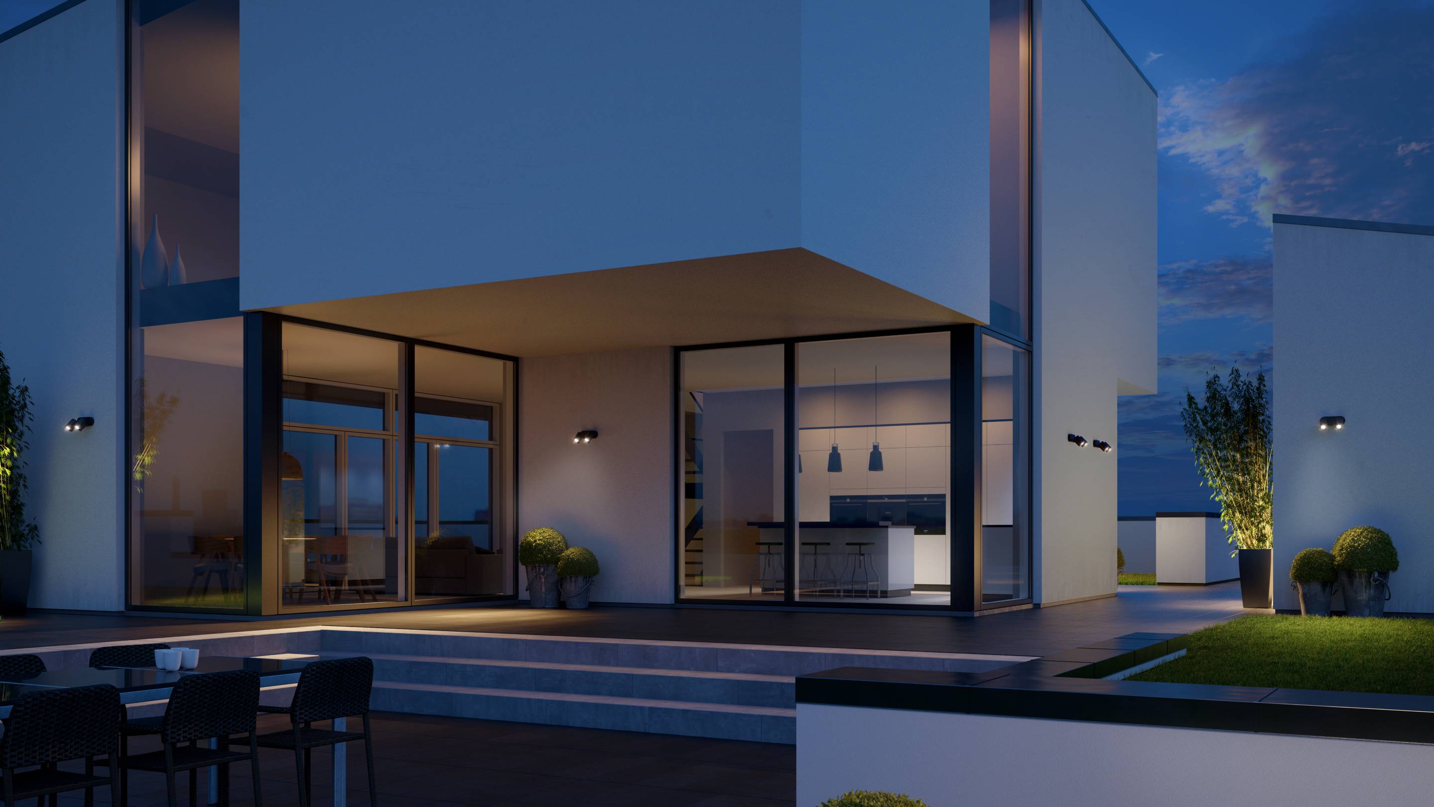 Modern verlicht huis 's nachts met grote glasgevels en terras. In het interieur zijn keukenonderdelen en hangende verlichting zichtbaar.