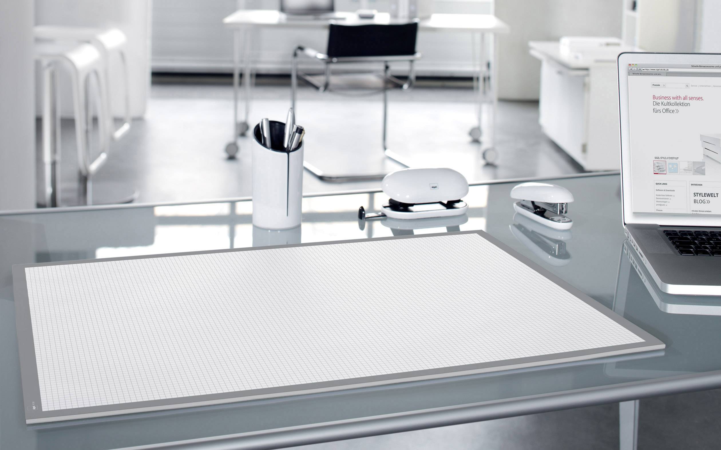 Moderne werkplek met een transparante glazen tafel, waarop een laptop, een bureauonderlegger, een pennenhouder, een perforator en een muis. Lichte, minimalistische omgeving.