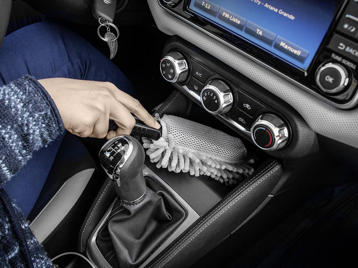 Hand reinigt bedieningspaneel van de airconditioning in een auto met een grijs stofdoek. Draaiknoppen en radioscherm zijn zichtbaar.