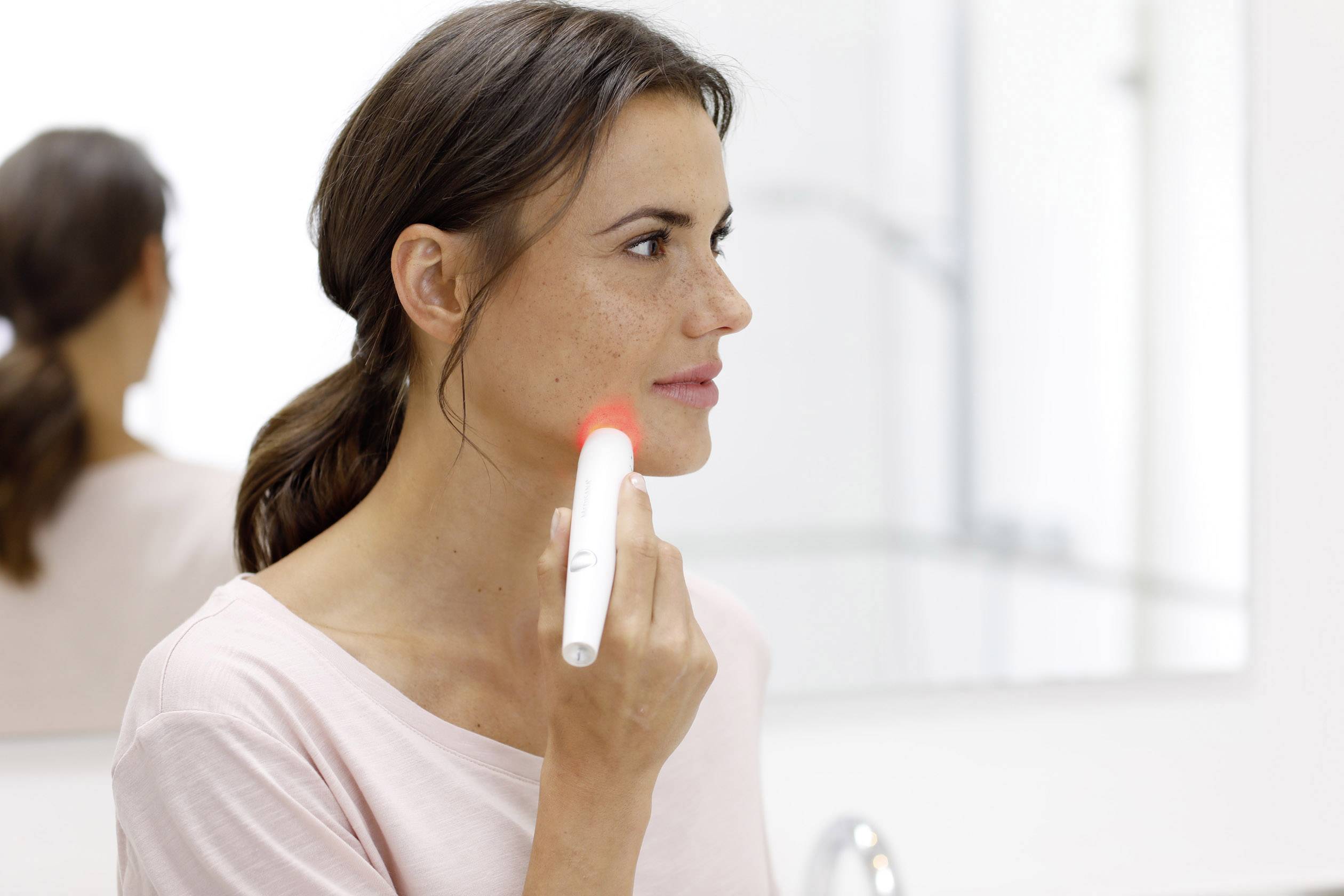 Een vrouw gebruikt een handapparaat met rood licht op haar gezicht. Ze lijkt het zacht tegen haar wang te drukken.