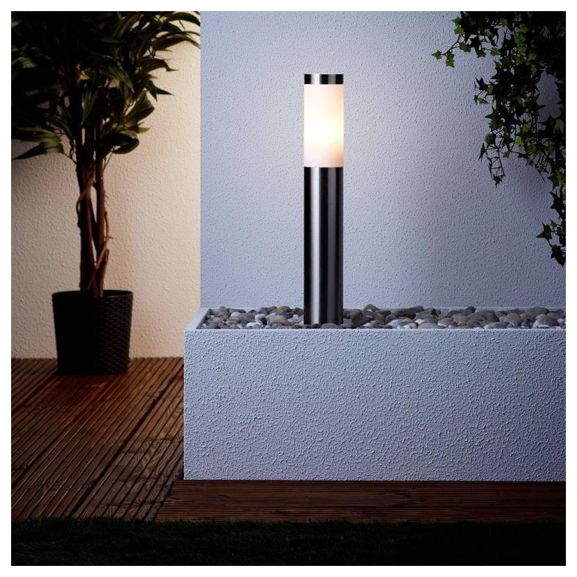 Een moderne buitentuinlamp met een strak metalen ontwerp staat verlicht in een met kiezels gevulde hoek naast een potplant onder zacht licht.