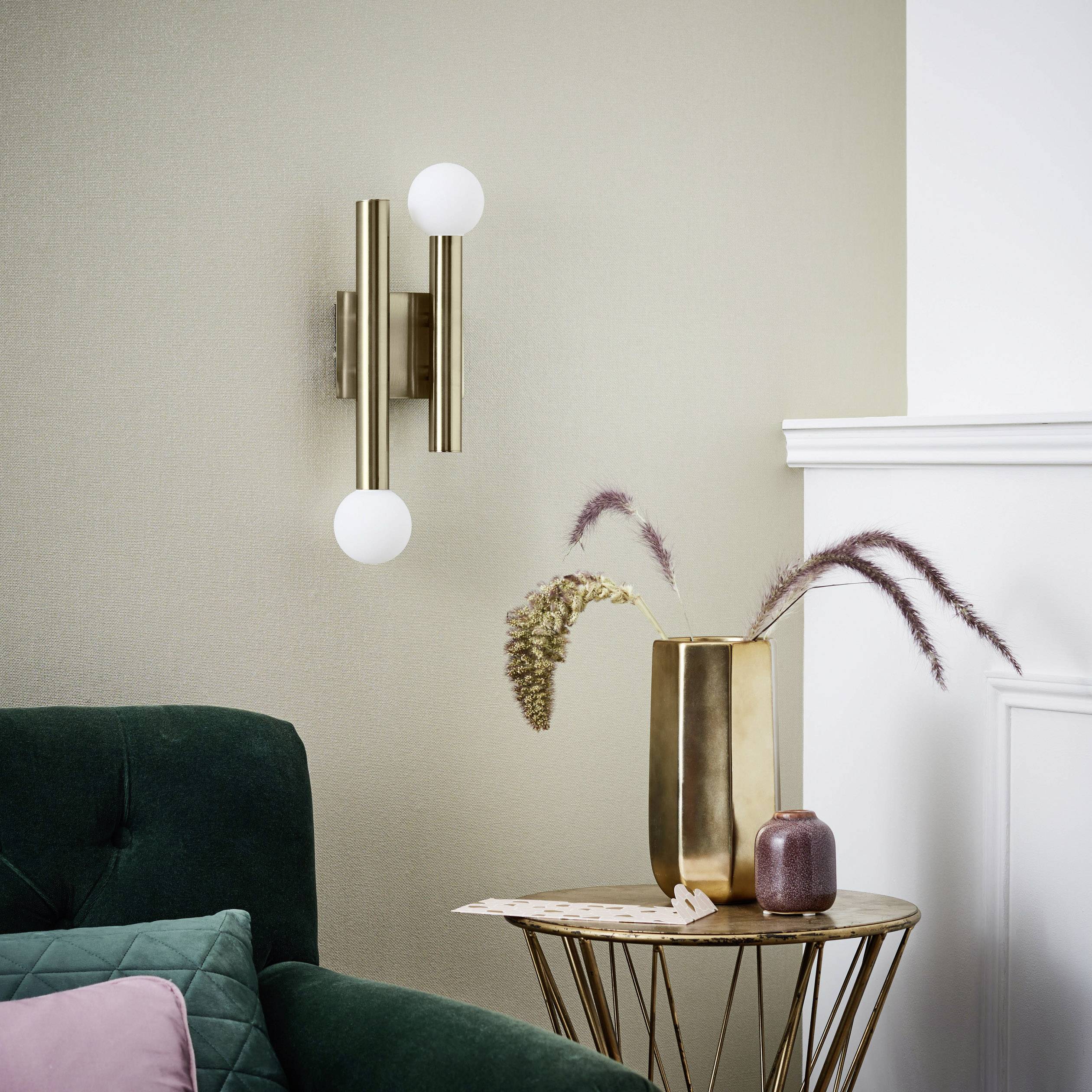 Wandlamp in retro-stijl met twee ronde armaturen, naast een groene bank en gouden vaas op een bijzettafel. Woonkamersfeer.