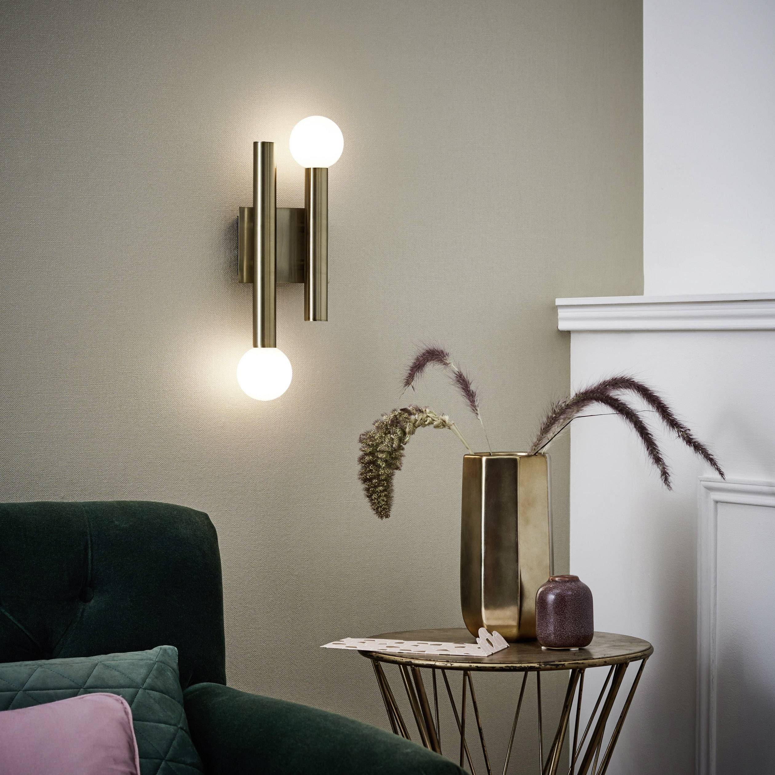 Wandlamp met twee bol-vormige lampen aan een woonkamermuur. Ernaast een groene fauteuil en een ronde tafel met vaas en decoratieve elementen.