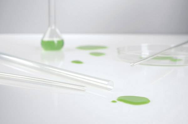 Een omgevallen reageerbuis met groene vloeistof op een laboratoriumtafel, met andere laboratoriumapparatuur op de achtergrond.