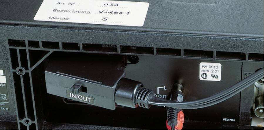 'Achterkant van een elektronisch apparaat, zwarte kabel aangesloten met IN/OUT-aanduiding, bovenaan een etiket met opschrift Video1.'
