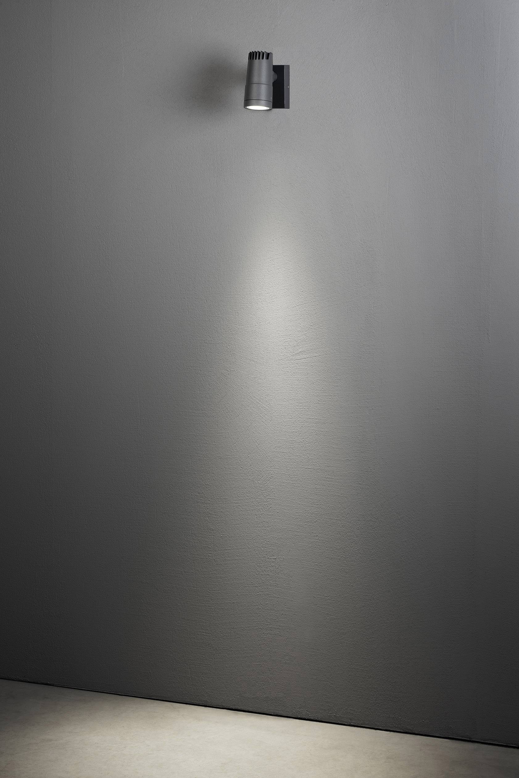 Een zwarte wandlamp schijnt op een sobere grijze muur en creëert een lichtkegel die op een gedeeltelijk verlicht grondvlak valt.