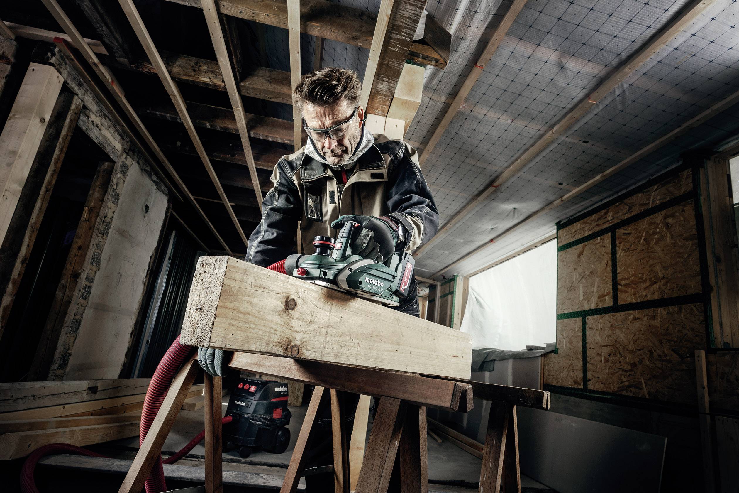 Een man werkt met een cirkelzaag in een houten werkplaats. Hij draagt een veiligheidsbril en gehoorbescherming.