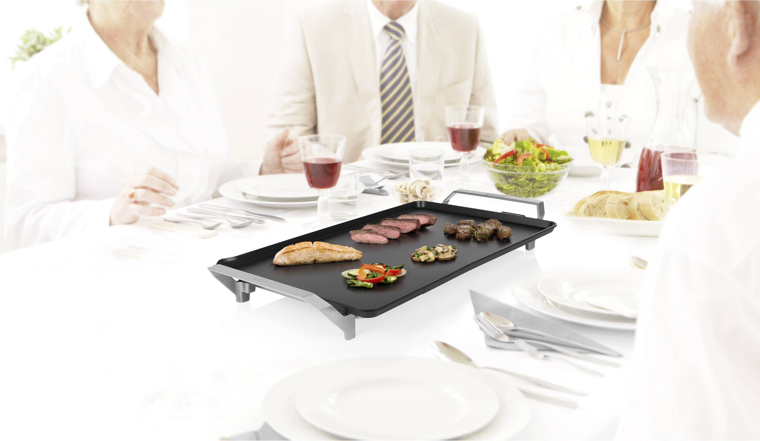 Een tafel waar vier personen in het wit gekleed zitten. In het midden staat een elektrische tafelgrill met verschillende gegrilde gerechten, ernaast een salade en dranken.