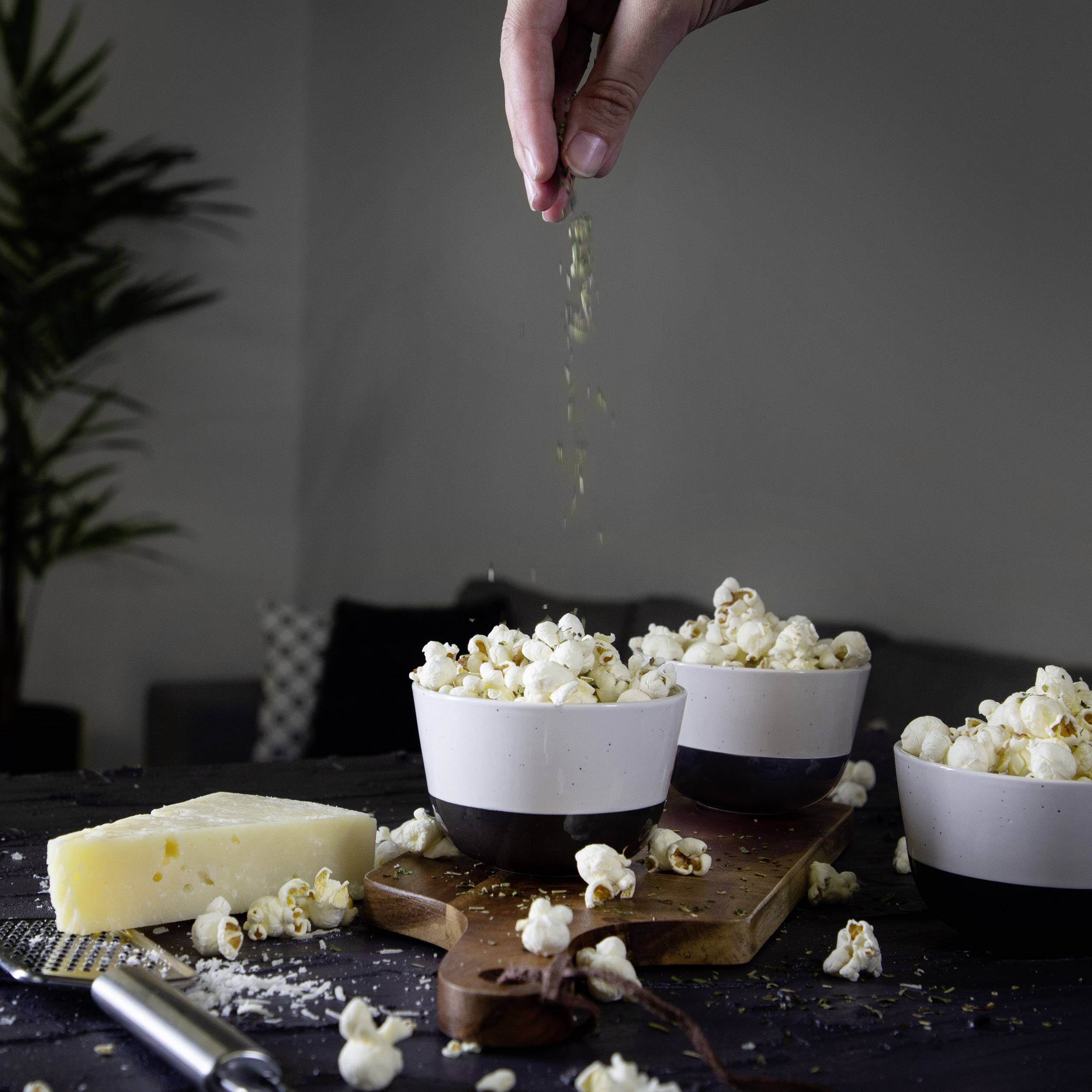 Een hand strooit kruiden over popcorn in witte kommen op een tafel met kaas en een plant op de achtergrond.