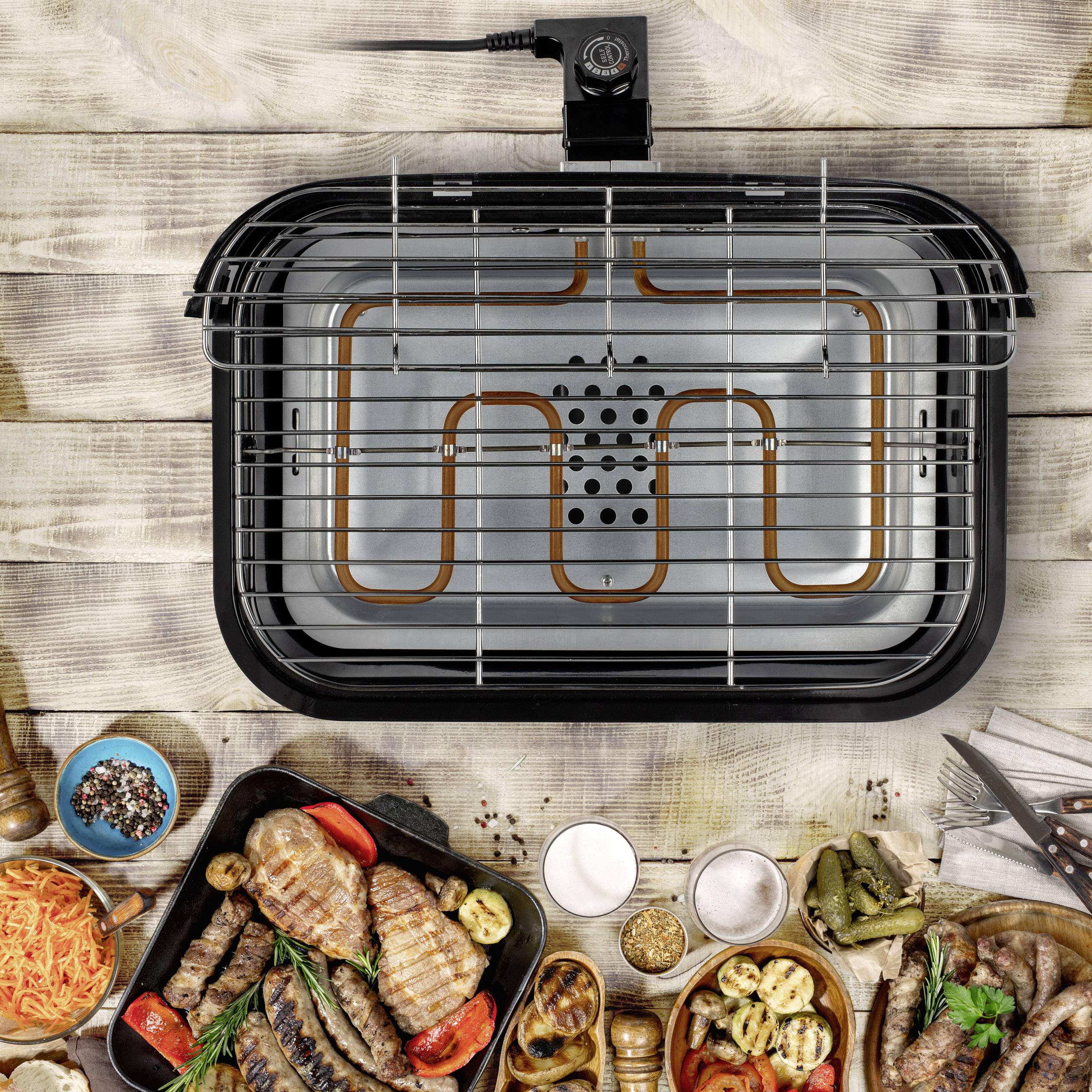 Elektrische grill met verwarmingselement van bovenaf, omgeven door gegrild vlees, groenten en kruiden op een houten tafel.