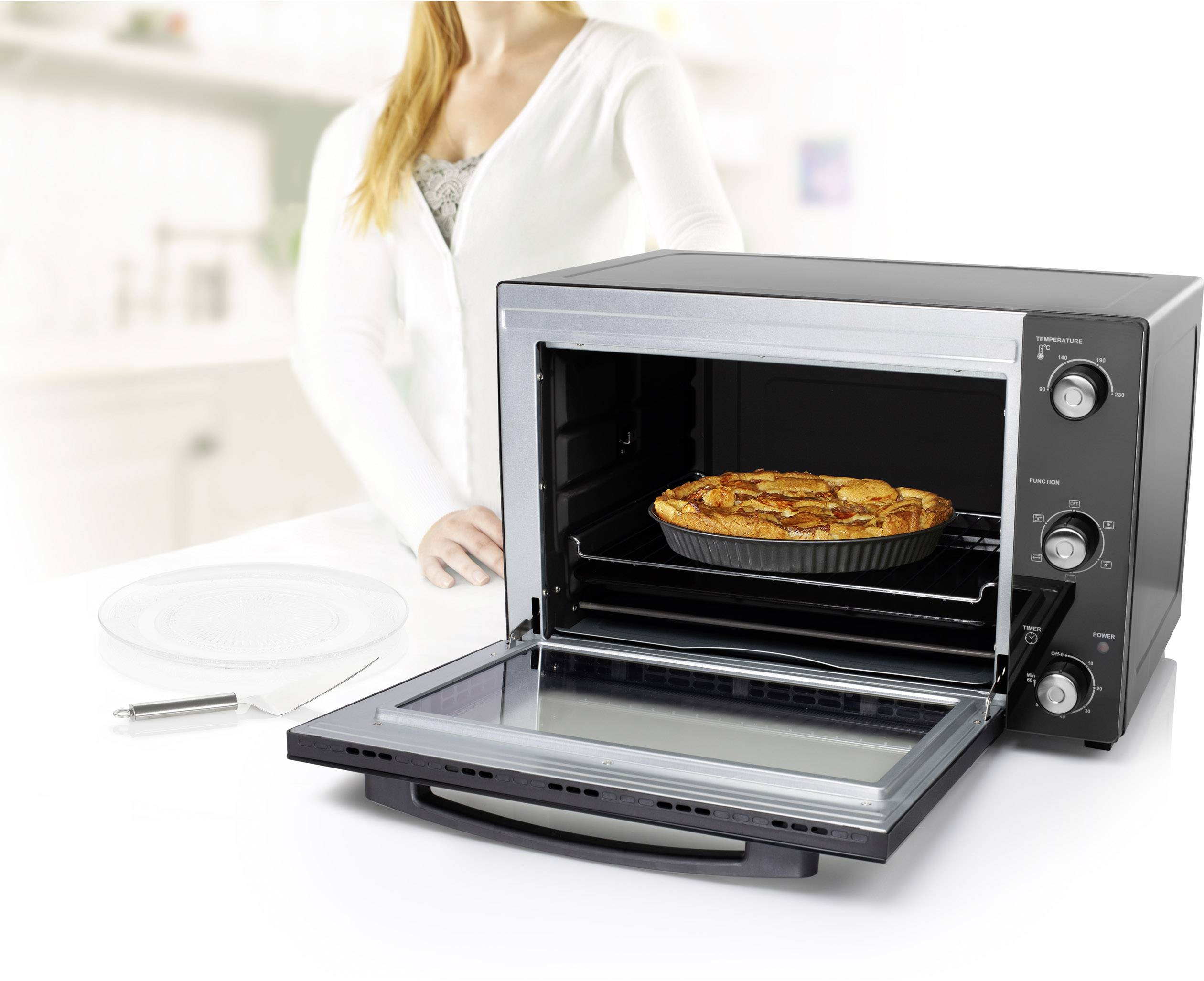 Een vrouw staat naast een geopende mini-oven, waarin een afgebakken cake op een rooster ligt.